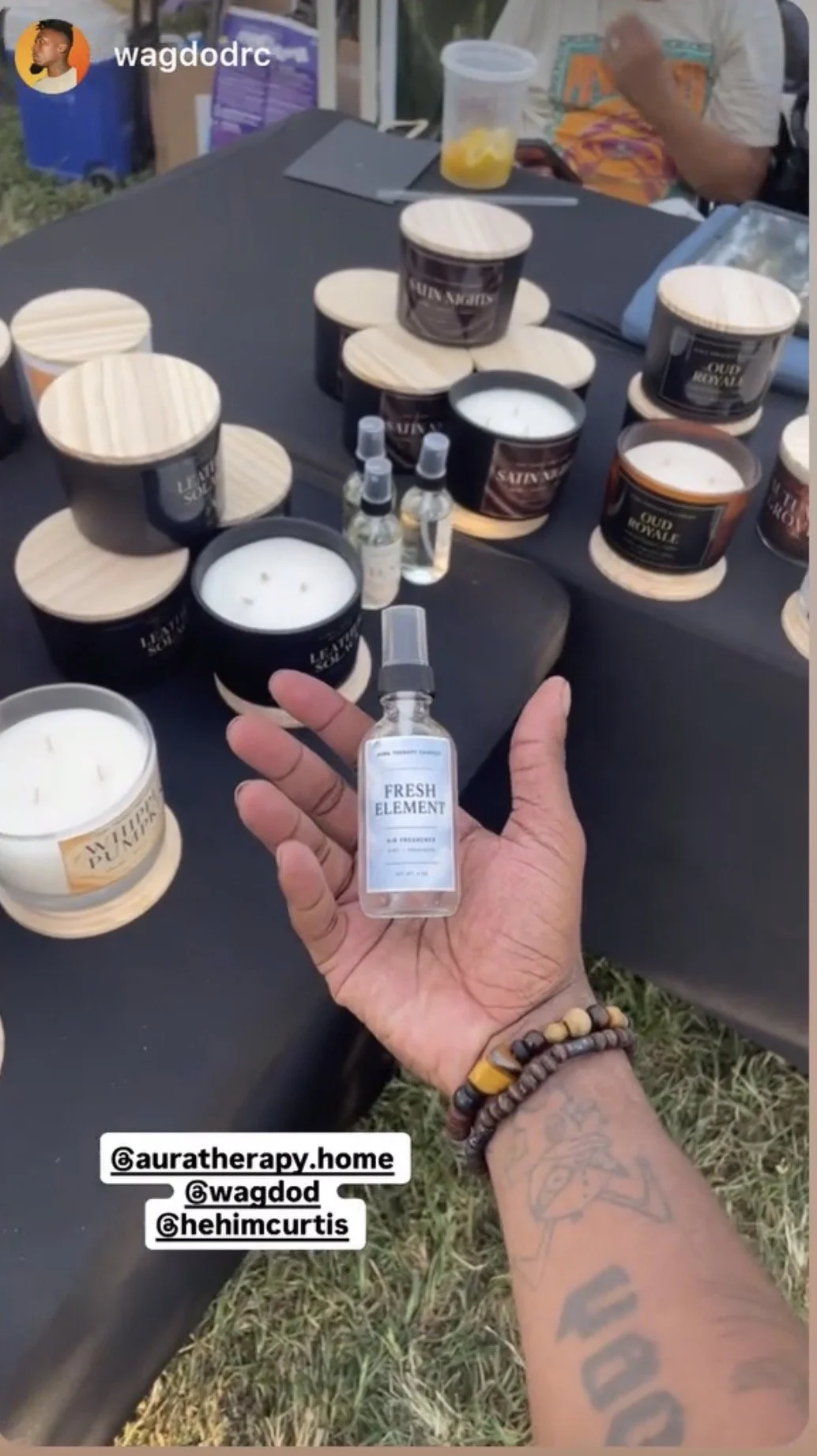 A person holding a spray bottle labeled "Fresh Element" in front of a table with black candles in wooden jars labeled "Lunar Soul" and "Old Divine," some with white candles inside. There are small spray bottles and a container of orange slices on the table. Another person is sitting behind the table with a drink and a tablet. The setting appears to be outdoors, possibly at a market or event.