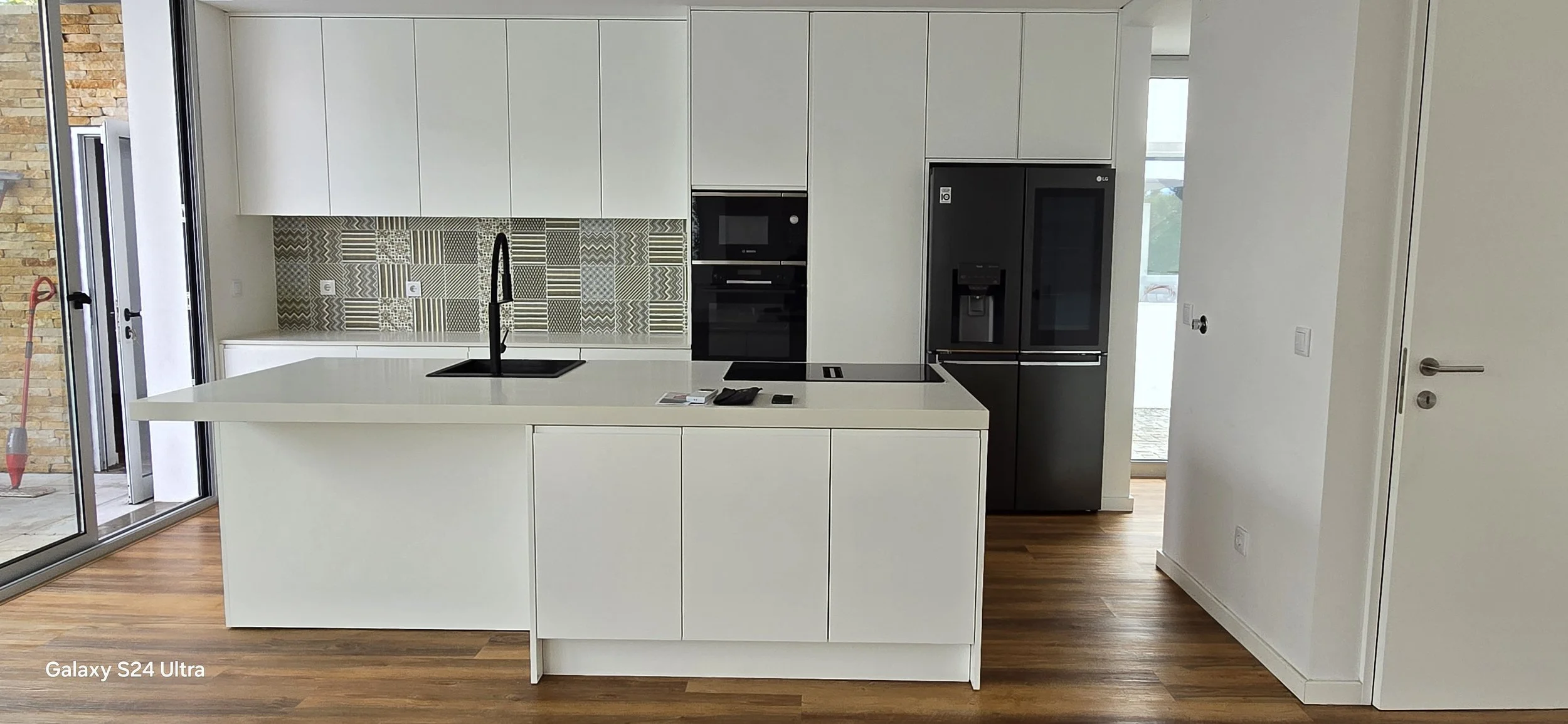 Cozinha moderna com armários brancos, bancada de ilha e eletrodomésticos embutidos, incluindo geladeira LG, fogão de indução, micro-ondas, e porta de vidro perto de uma parede revestida com azulejos decorativos em tons de cinza e branco.