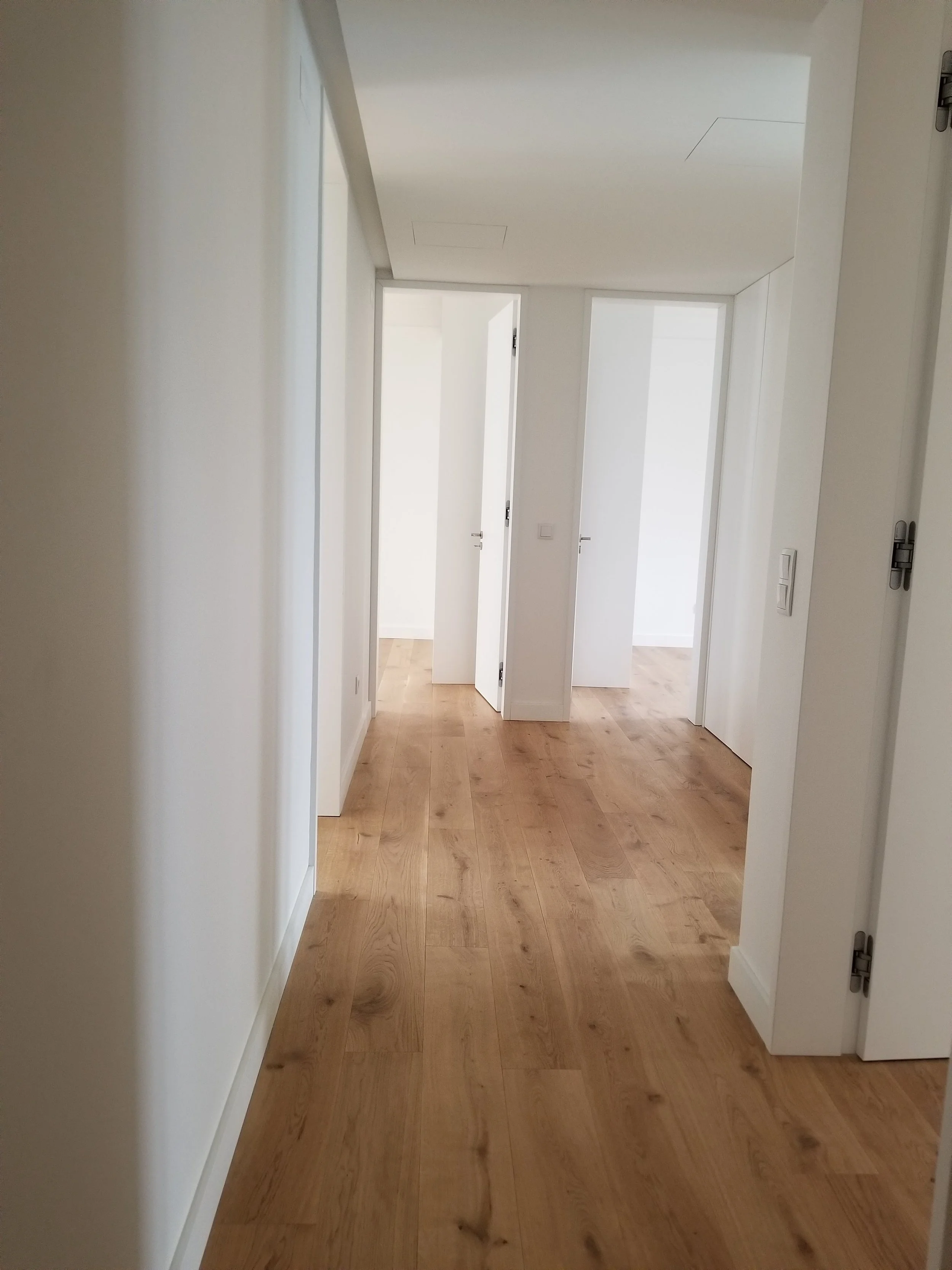 Corredor de uma casa com piso de madeira clara, paredes brancas e portas brancas até o teto, uma delas um pouco aberta.