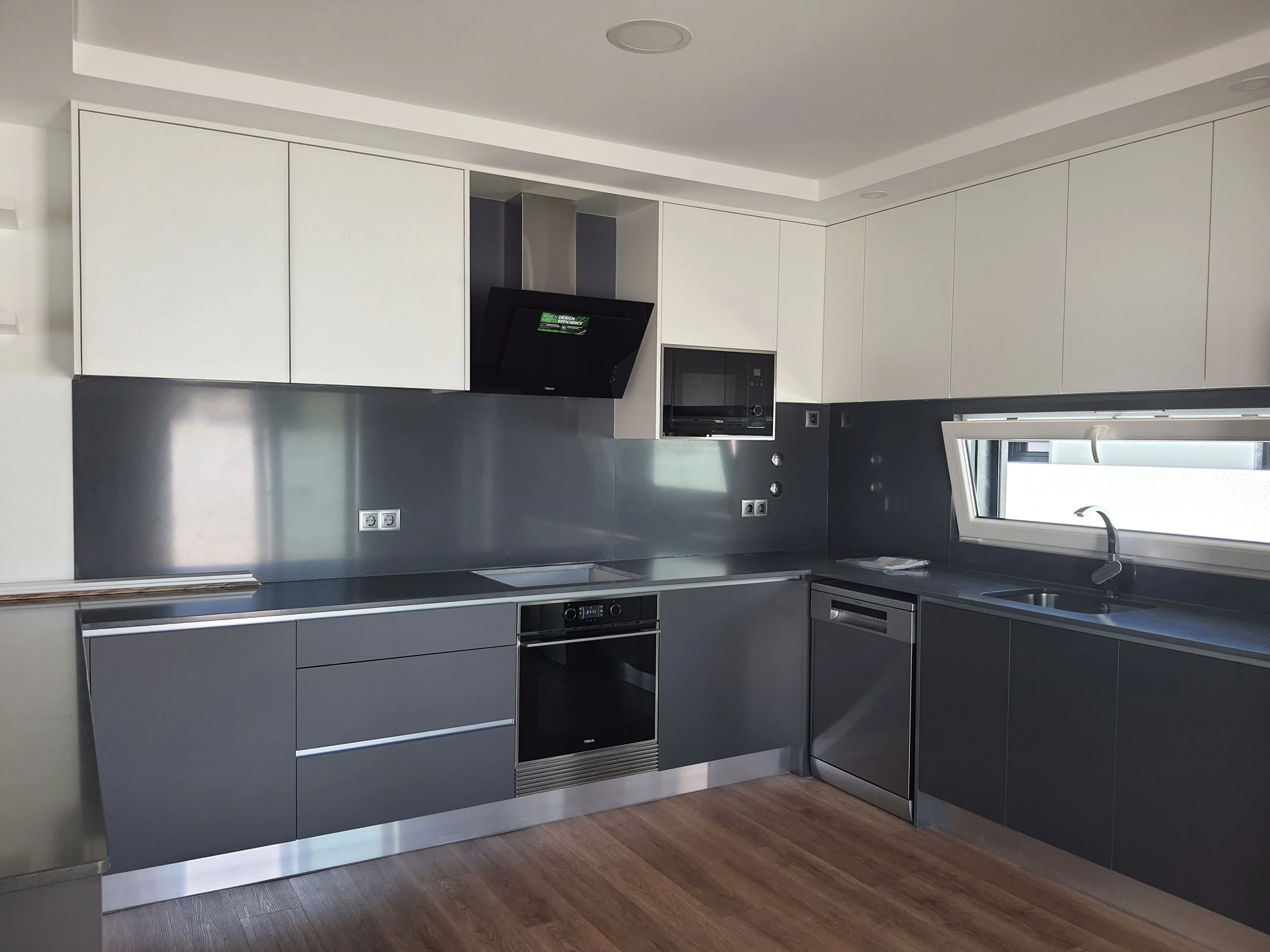 Cozinha moderna com armários brancos, bancada cinza escuro, eletrodomésticos embutidos e janela.