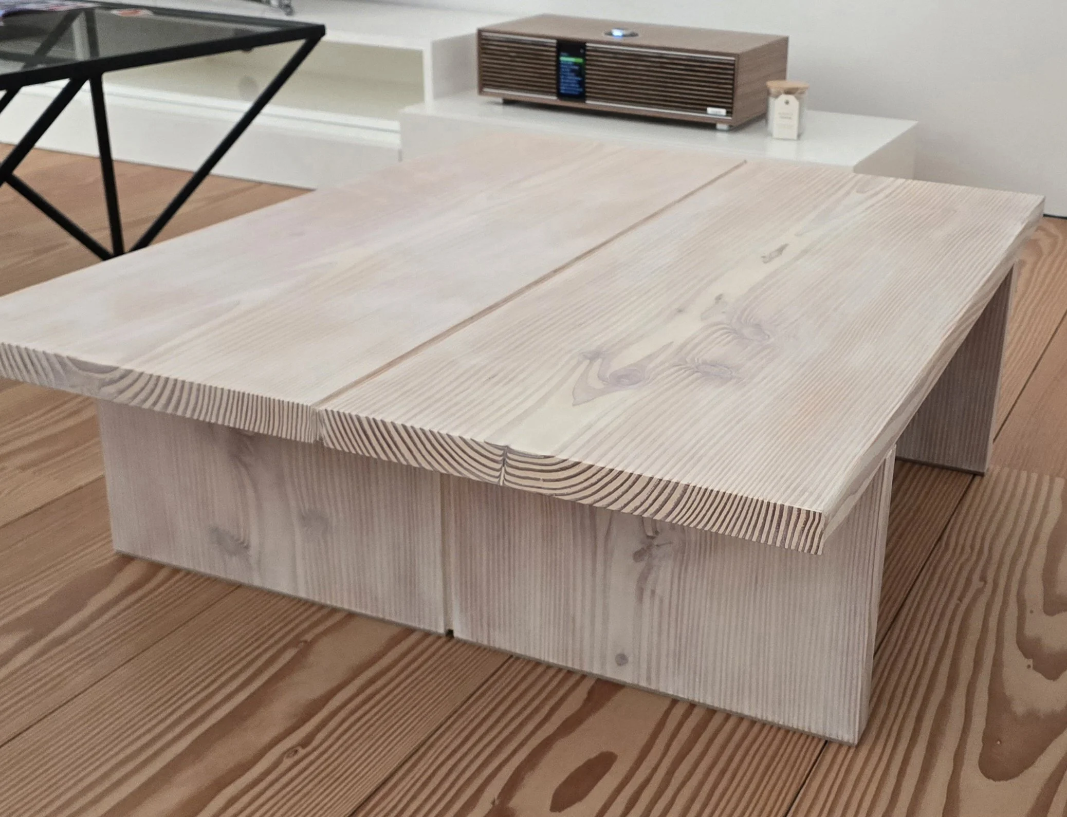 Mesa de madeira clara com revestimento de ripas na borda, com um aparelho de som de madeira escura no fundo e uma janela de vidro ao lado.