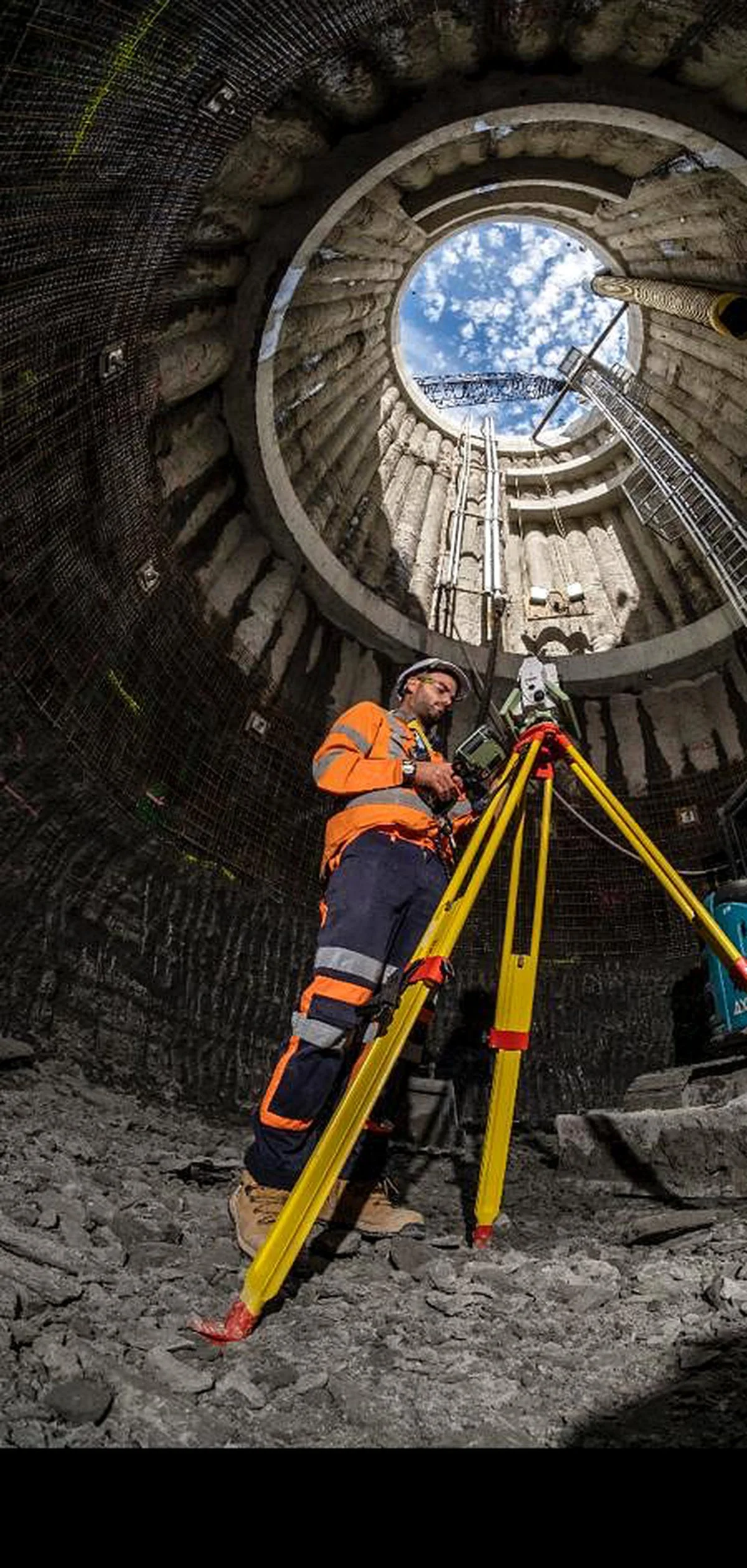 Construction worker in orange safety gear and helmet operating surveying equipment inside a large, circular stone structure with a view of a partly cloudy sky above.