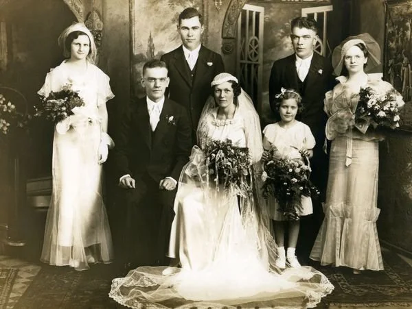 A black-and-white vintage wedding photograph showing seven people. The bride is seated in the center wearing a traditional wedding dress and veil, holding a large bouquet. The groom is to her left, wearing a dark suit and glasses. Three women, two holding large bouquets, and one young girl with a bouquet, stand around them. Two men are behind the bride and groom. The setting appears to be an ornate indoor space with decorative walls and a patterned rug.