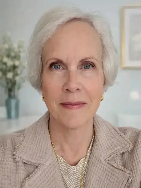 An elderly woman with short, white hair and blue eyes, wearing a beige jacket, gold jewelry, and a patterned top, standing in a bright, well-decorated room with a blurred background. Jewelry appraisal and heirloom jewelry collection