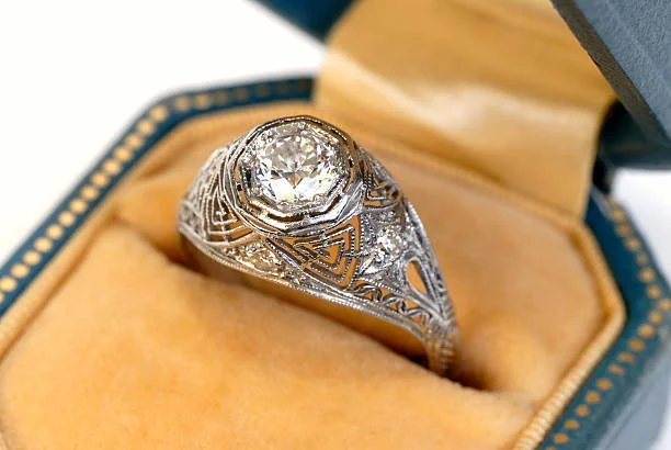 An ornate silver ring with a large central diamond in a jewelry box with a yellow interior.