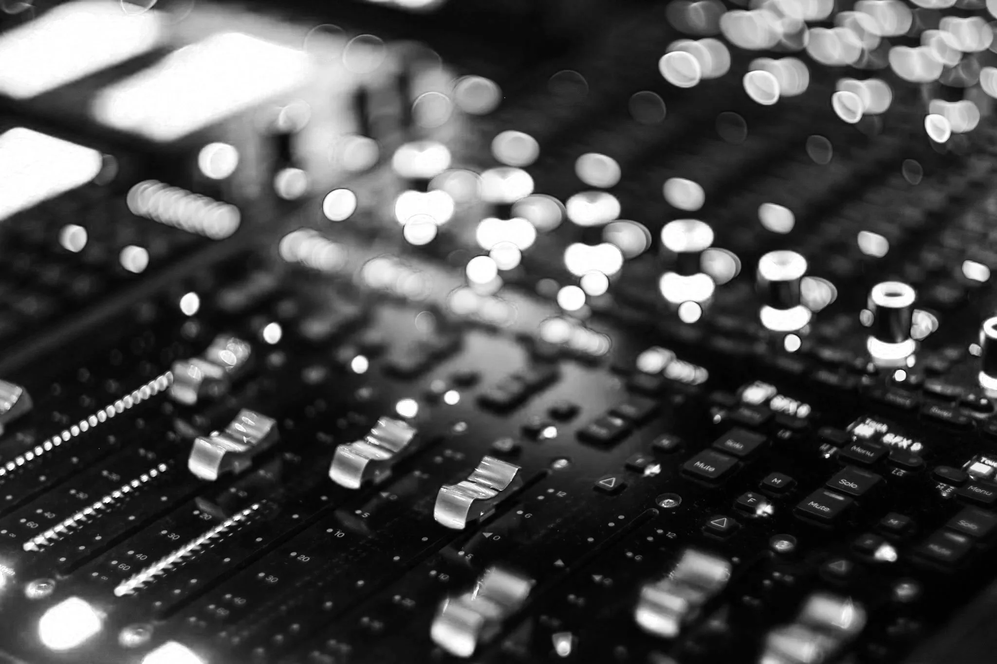 Close-up of an audio mixing console with blurred control knobs and sliders in black and white.