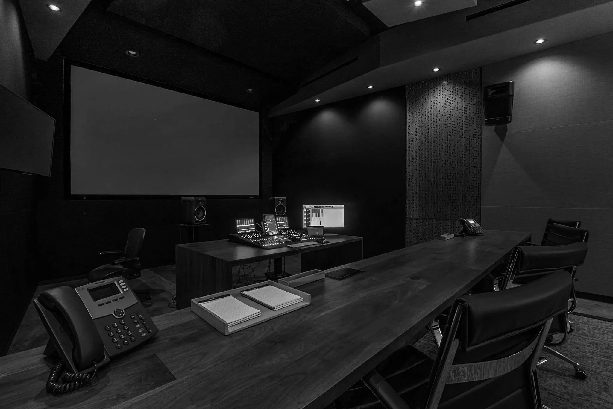 Dark industrial recording studio with black walls and ceiling, large screen on the wall, multiple audio control consoles, computers, and office chairs. Several telephones are on the desks.