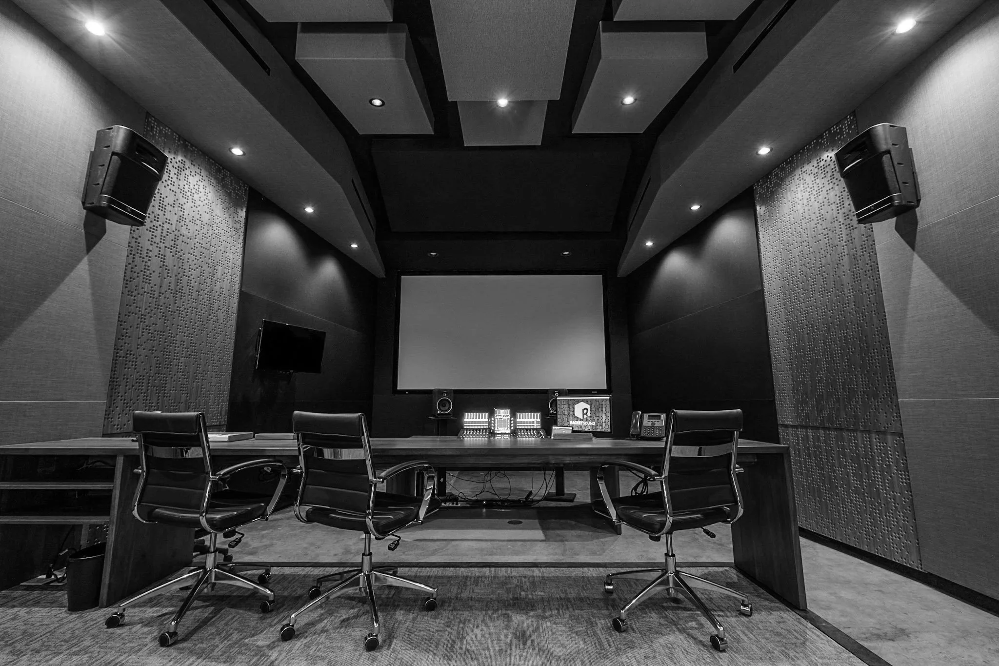 A professional sound mixing and recording studio with a large mixing console, multiple chairs, a large screen, two speakers mounted on the walls, and acoustic panels on the ceiling.