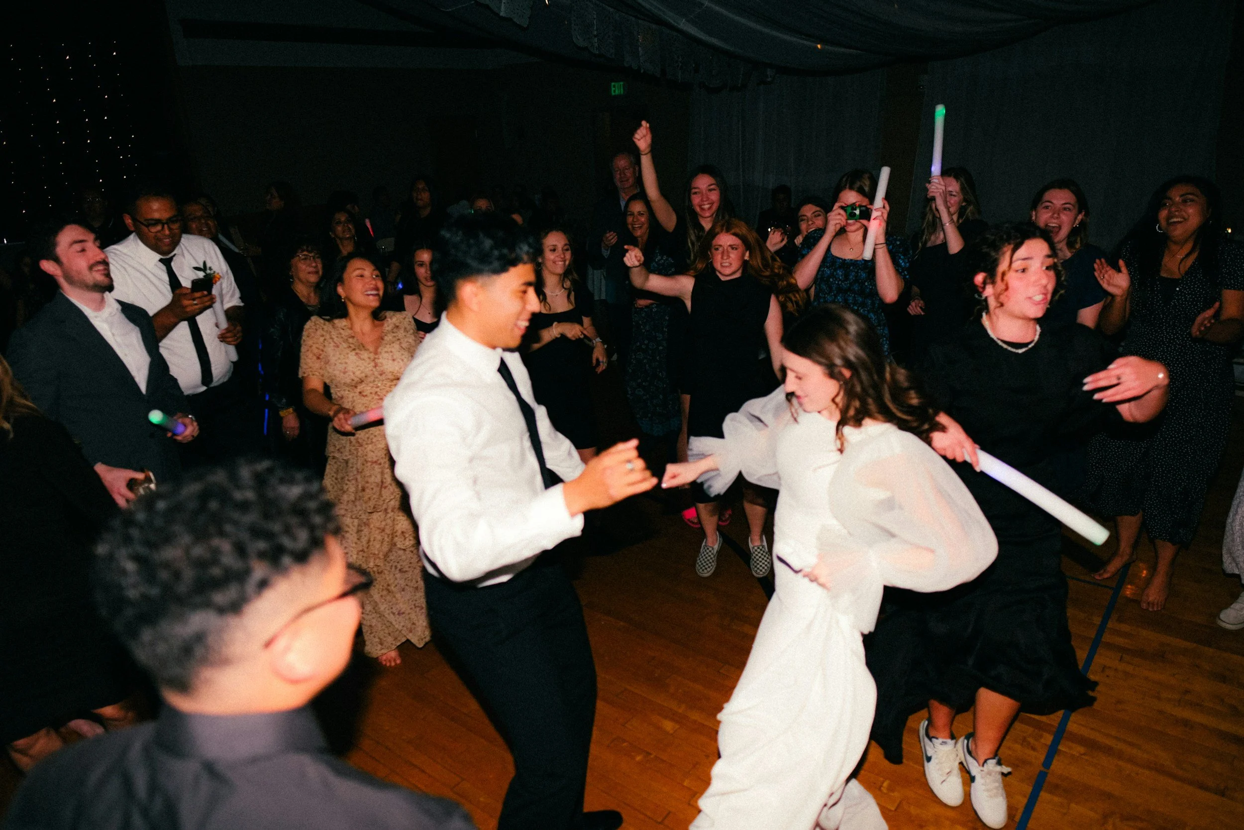 A group of people at a party or dance event, some dancing and others watching, with joyful expressions, in a dimly lit room with a wooden floor and dark curtains.
