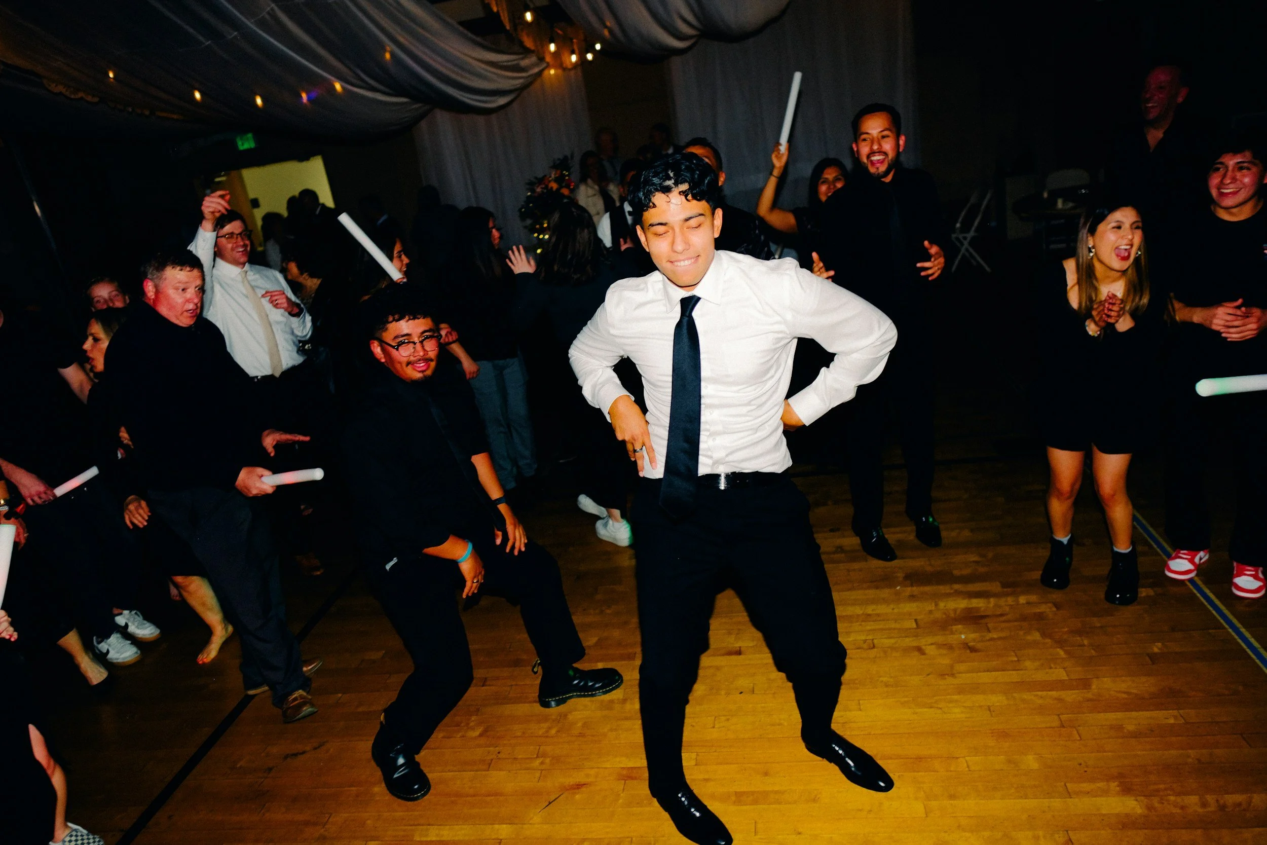 Group of people dancing and enjoying themselves at a party or celebration with a man in a white shirt and black tie in the foreground, some holding glow sticks, in a dimly lit venue.