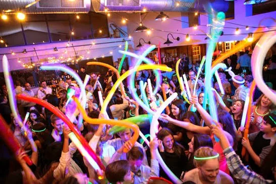 Crowd of people at a lively party or dance event with colorful glow sticks and LED lights, creating a festive atmosphere.