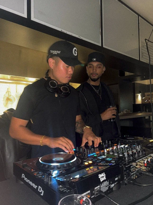 Two men DJing at a club or party, one wearing a black cap, headphones around neck, and gold bracelet; the other holding a drink, both operating DJ equipment.