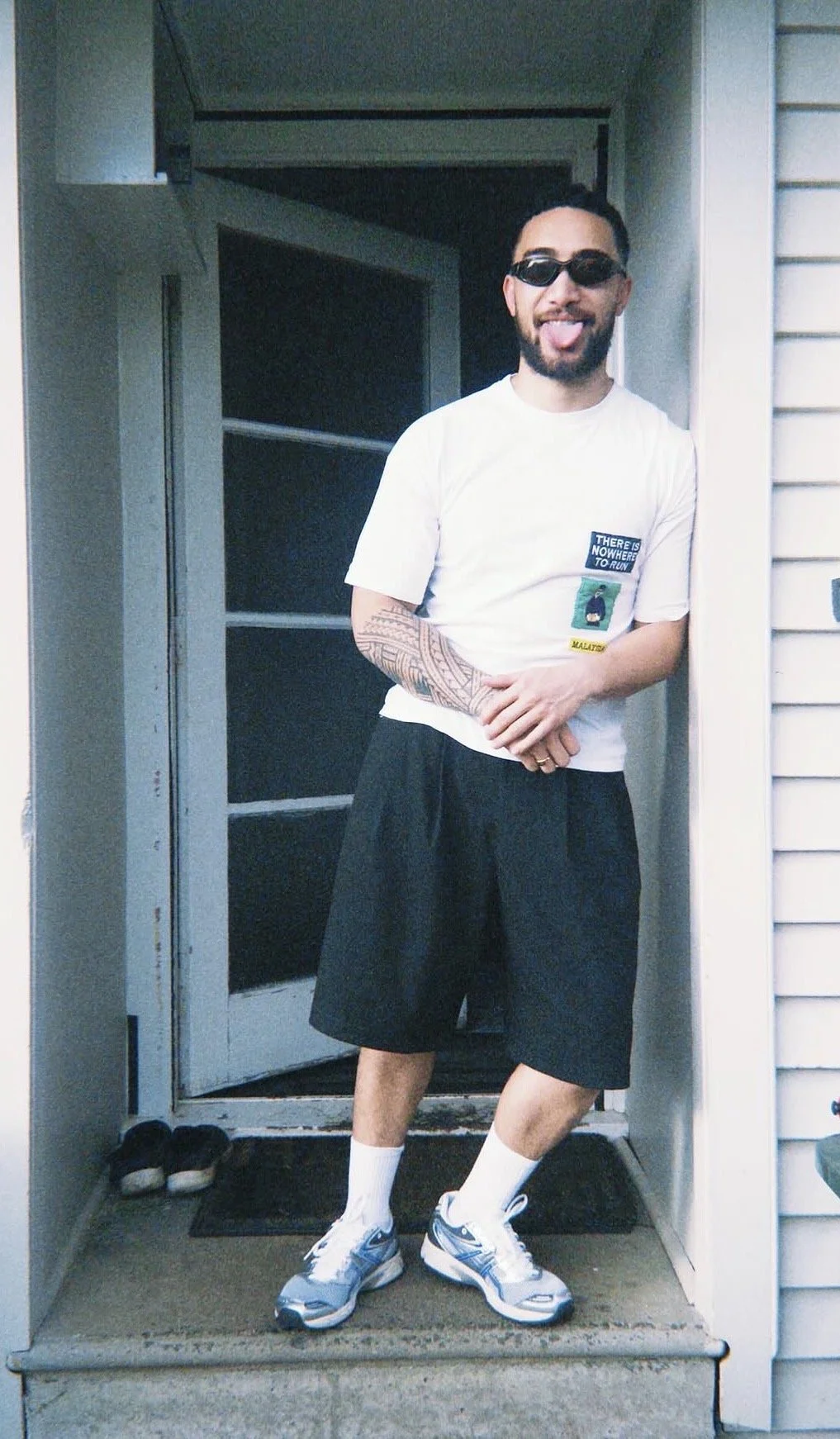 A man with a beard and sunglasses standing on a porch in front of a door, sticking his tongue out, wearing a white t-shirt with patches, black shorts, white socks, and sneakers.