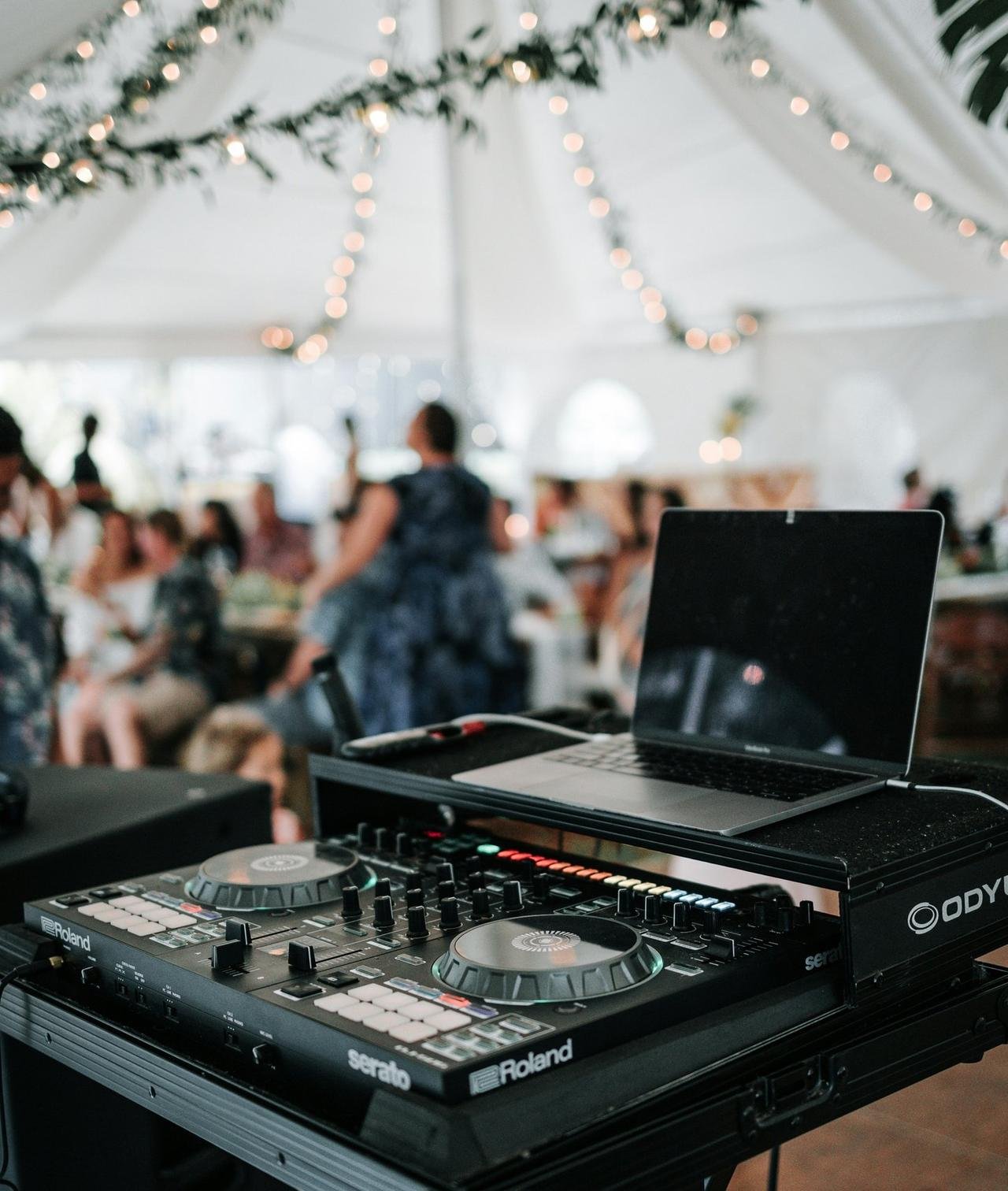 DJ equipment with a laptop on top, set up for a music event inside a venue decorated with string lights and hanging greenery, with people socializing in the background.