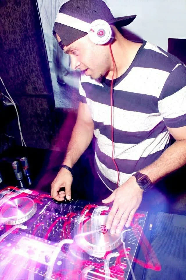 A young man wearing a striped t-shirt, a cap, and headphones is DJing with a professional turntable and mixer, focusing on the music.