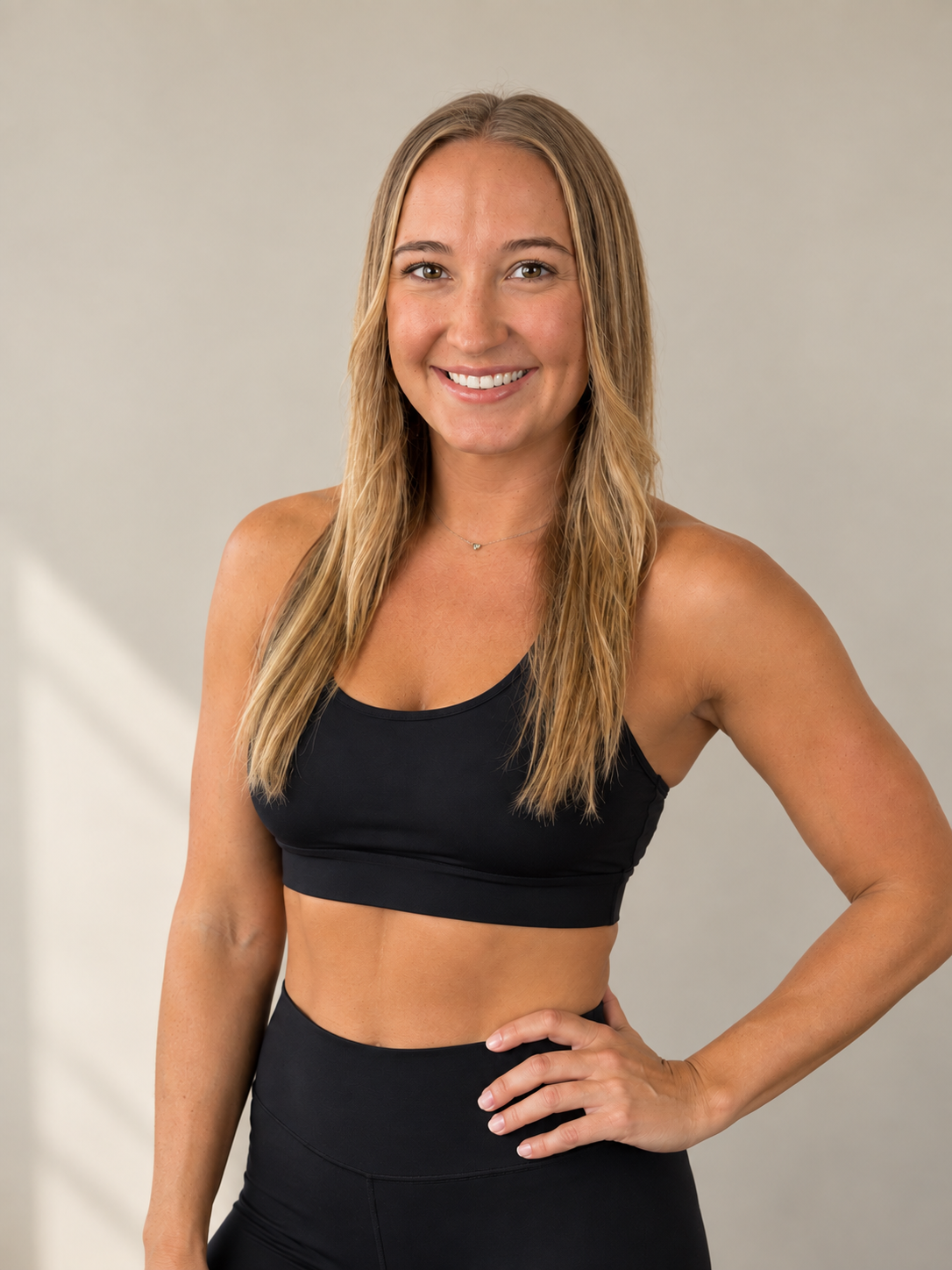 A smiling woman with long, wavy blonde hair wearing a black sports bra and black high-waisted workout leggings, standing against a plain light-colored background.