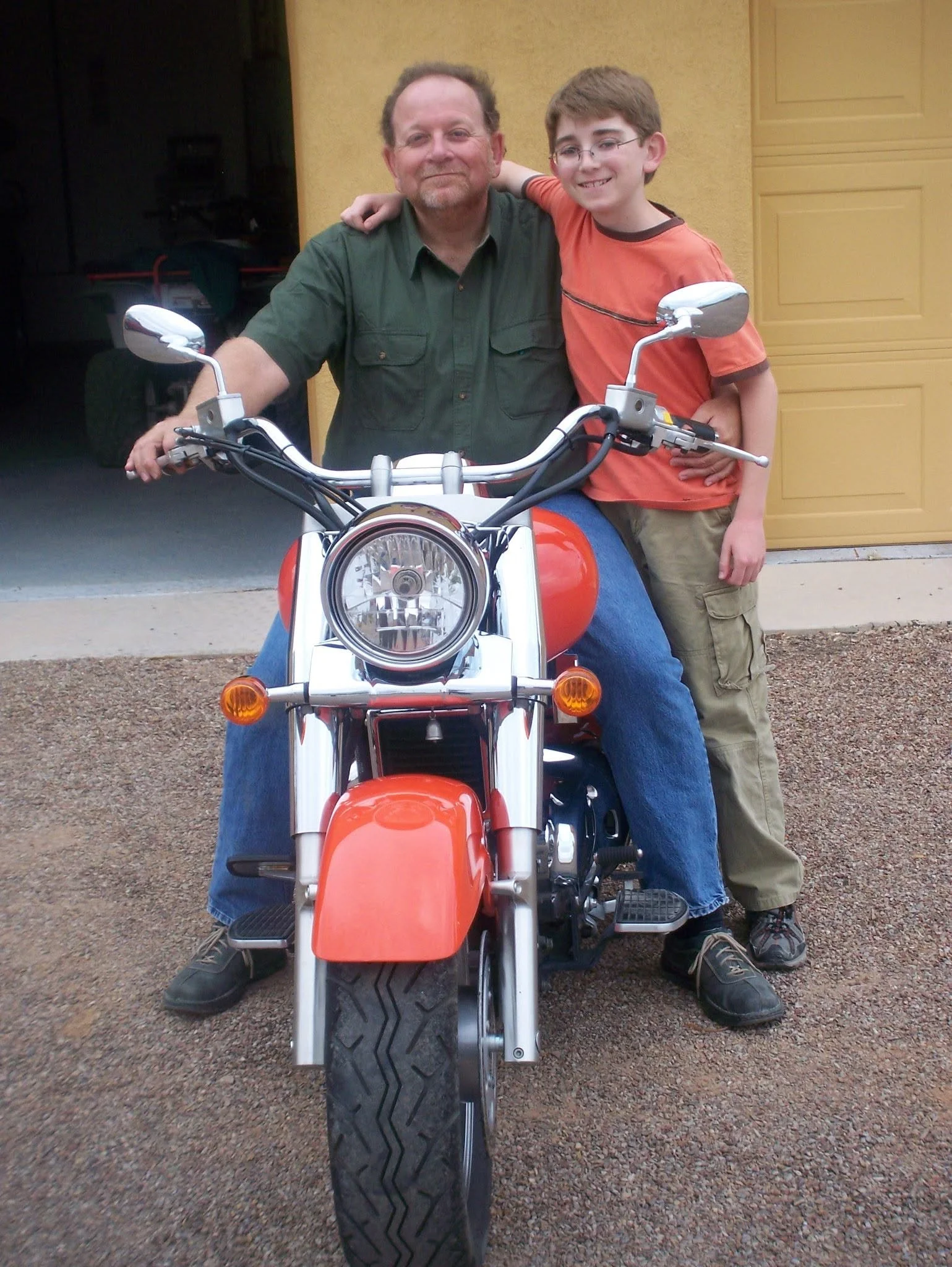 A man and a young boy standing together, with the boy's arm around the man's shoulder, next to a motorcycle in front of a yellow garage door.