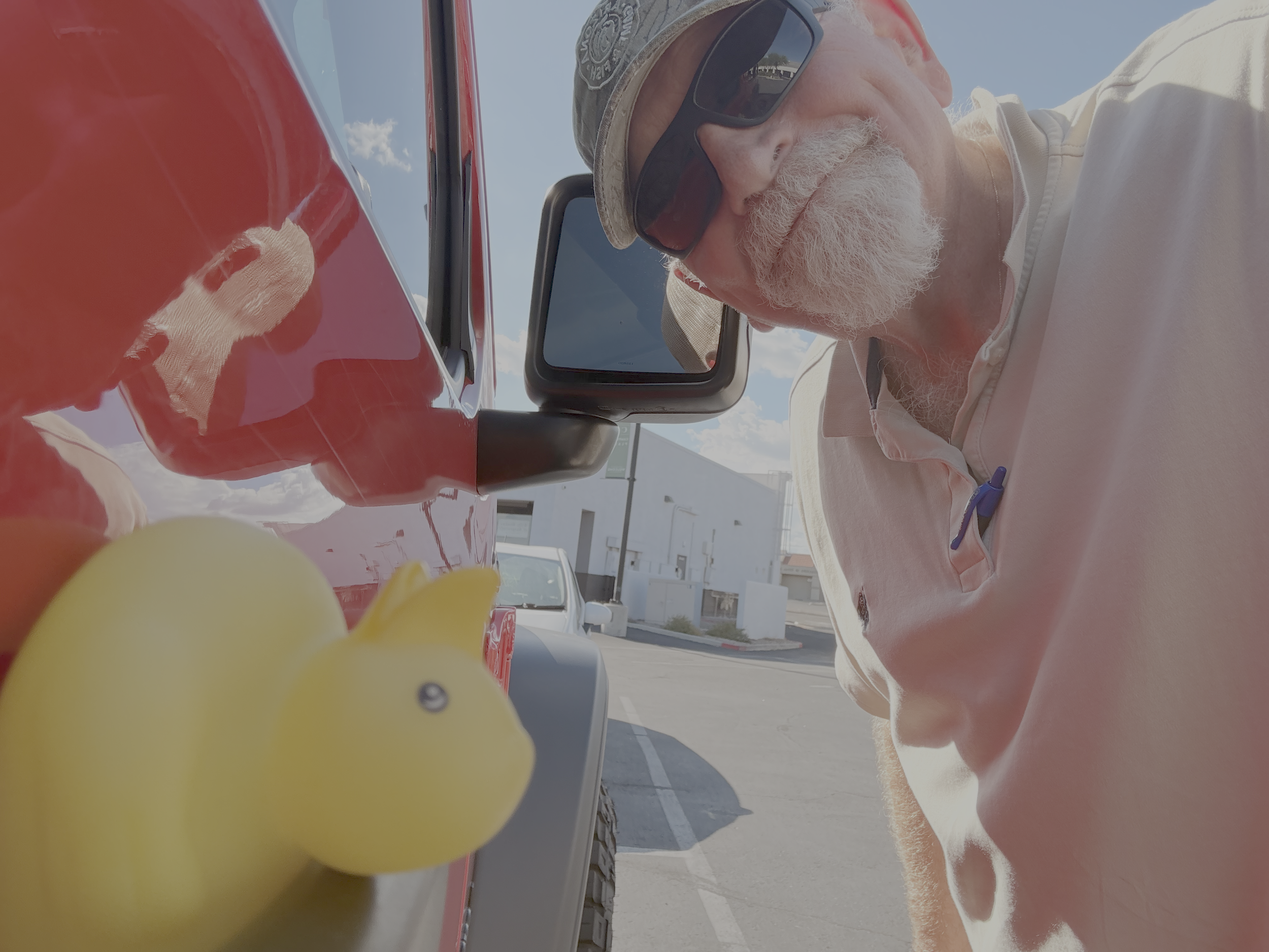 A man with a white beard and sunglasses wearing a beige cap and a light-colored polo shirt looking at a small yellow rubber duck on a car roof during daytime.