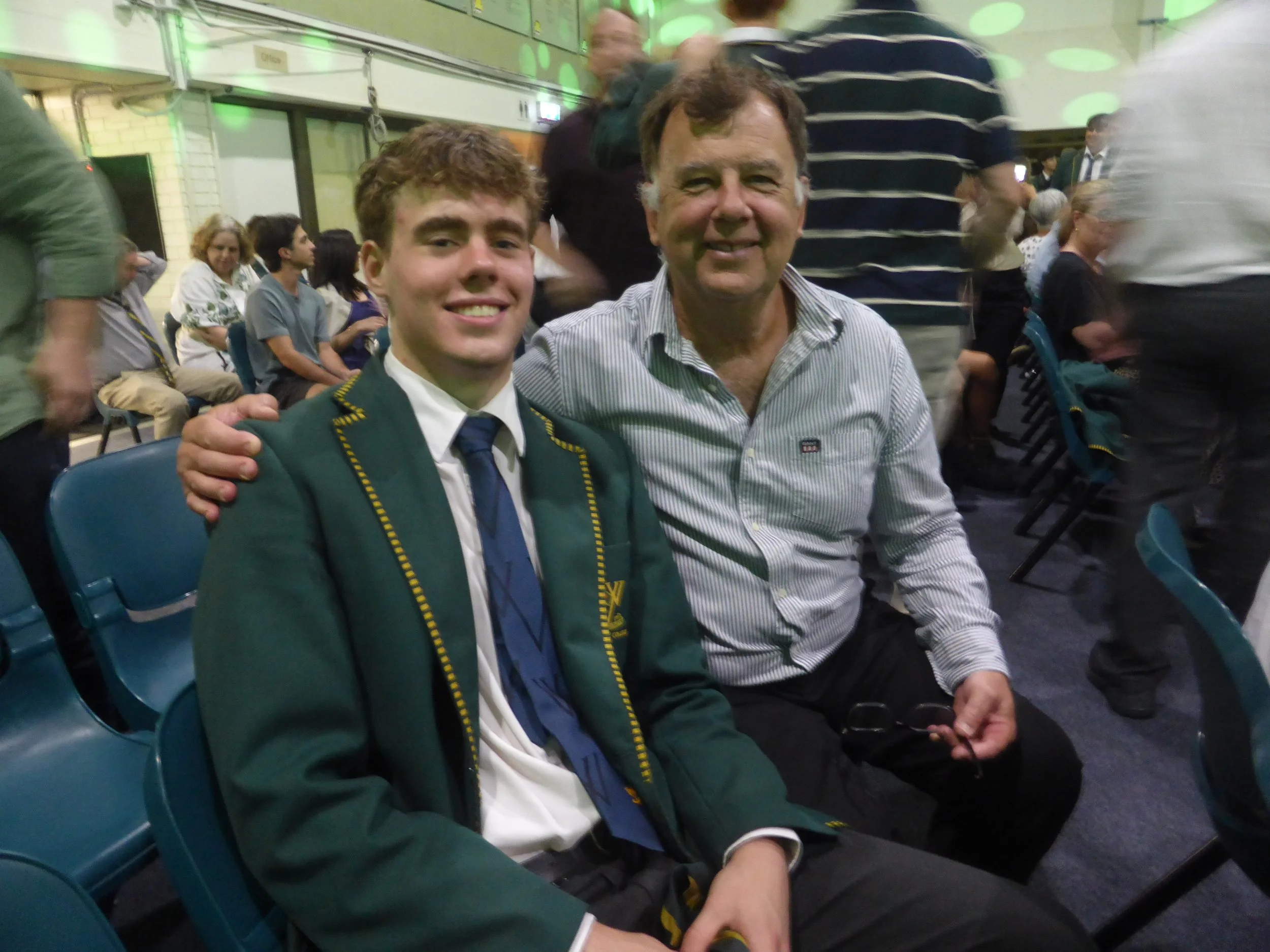 A young man in a school uniform sitting next to an older man, both smiling at the camera, in a crowded indoor event with green lighting and people seated in the background.