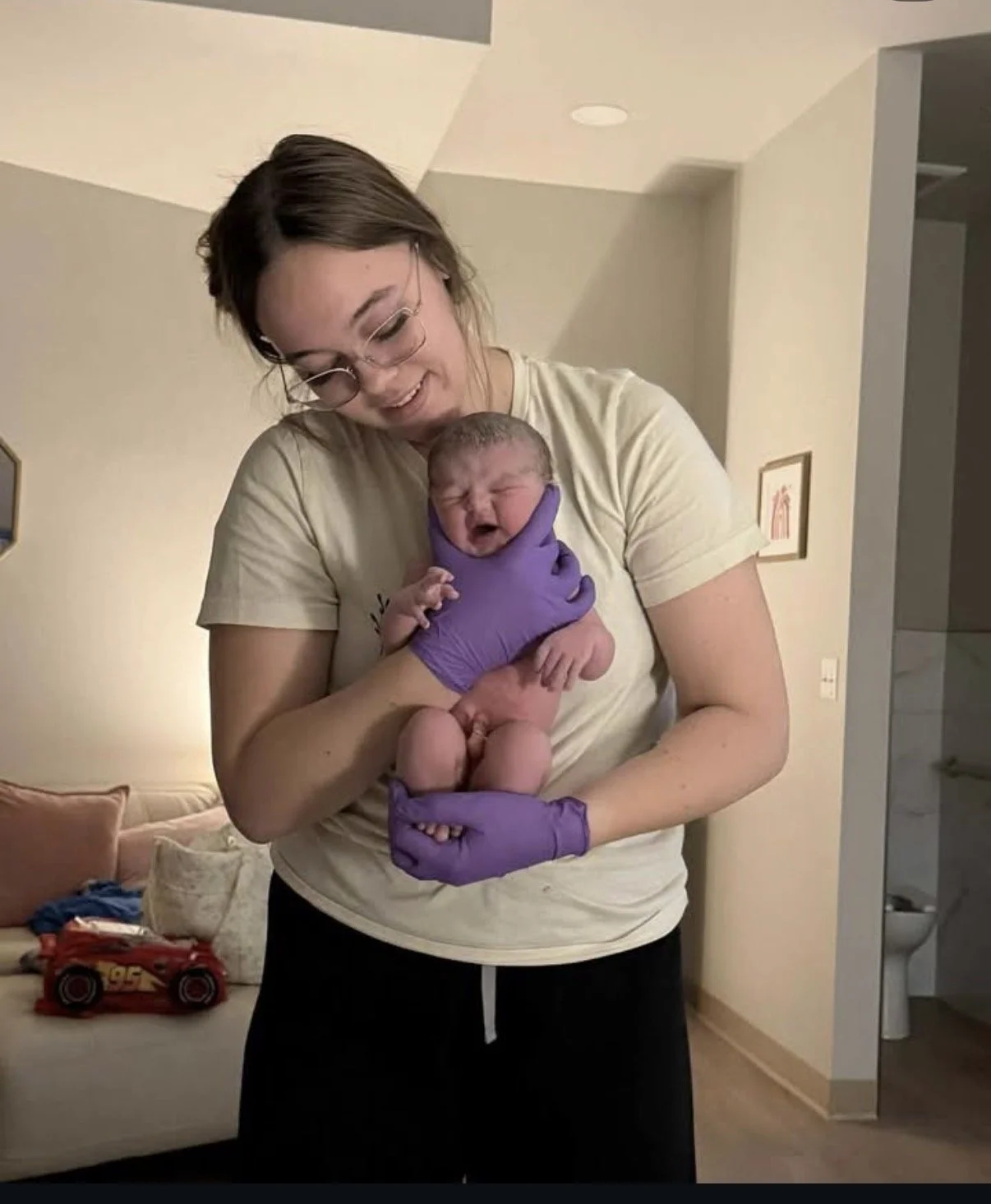 A woman with glasses and purple gloves holding a newborn baby. The woman is smiling and looking down at the baby, who is crying.