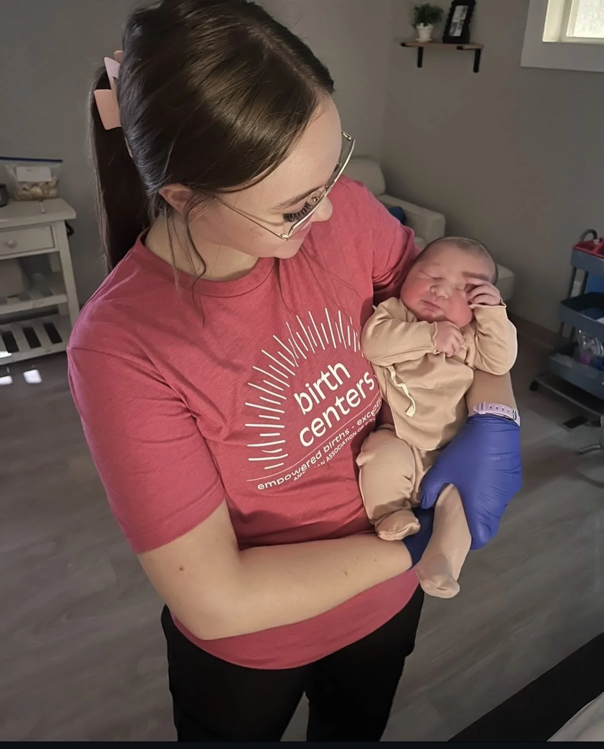 A woman holding a newborn baby in a hospital room. The woman is wearing a pink t-shirt with 'birth centers' printed on it and glasses. The baby is wrapped in beige clothing and resting against her chest.