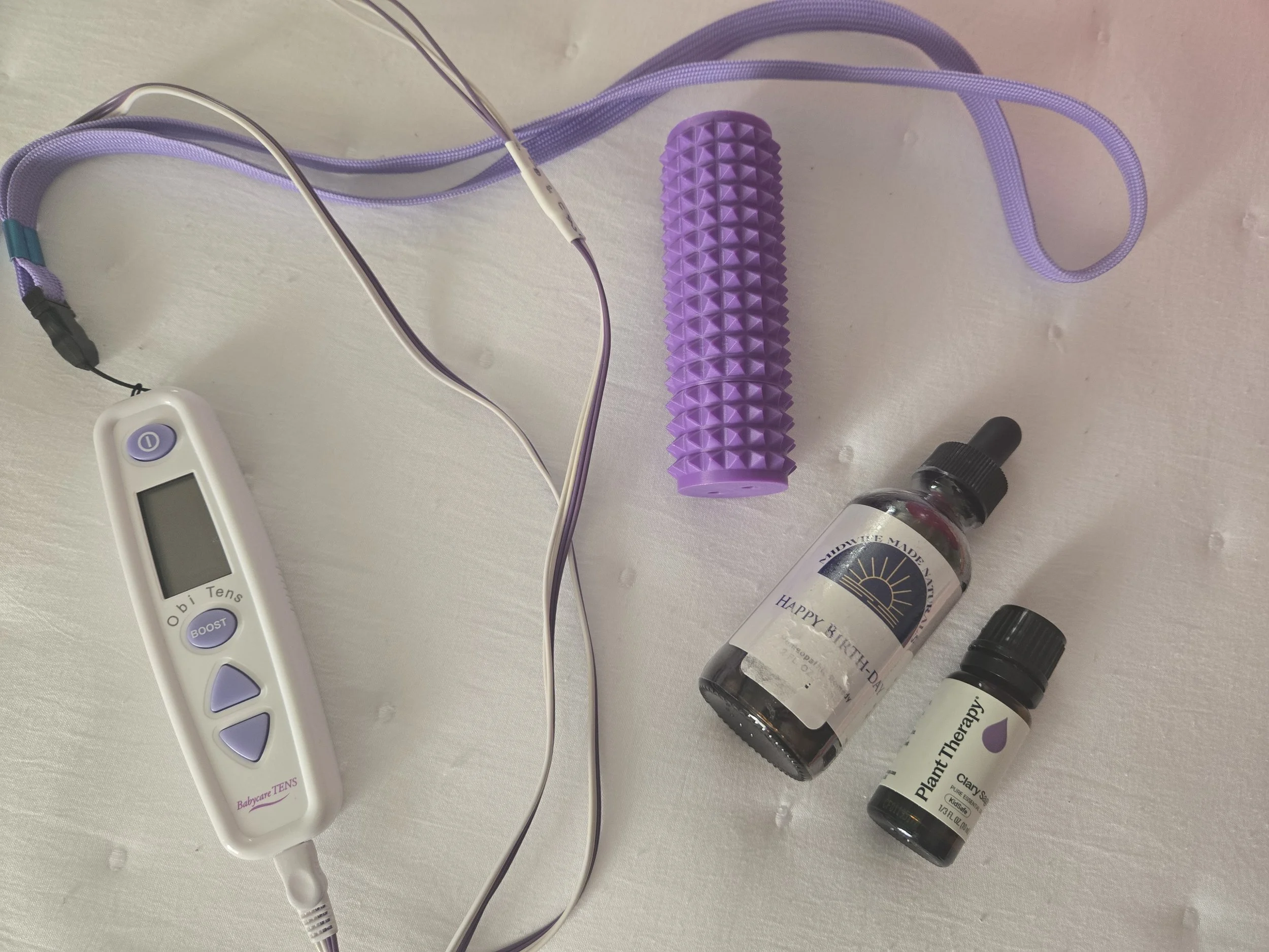 A collection of therapy and wellness items including a purple massage roller, a white digital thermometer, small bottles of plant therapy essential oils, and a purple and white vibration therapy device with wires, set on a white surface.