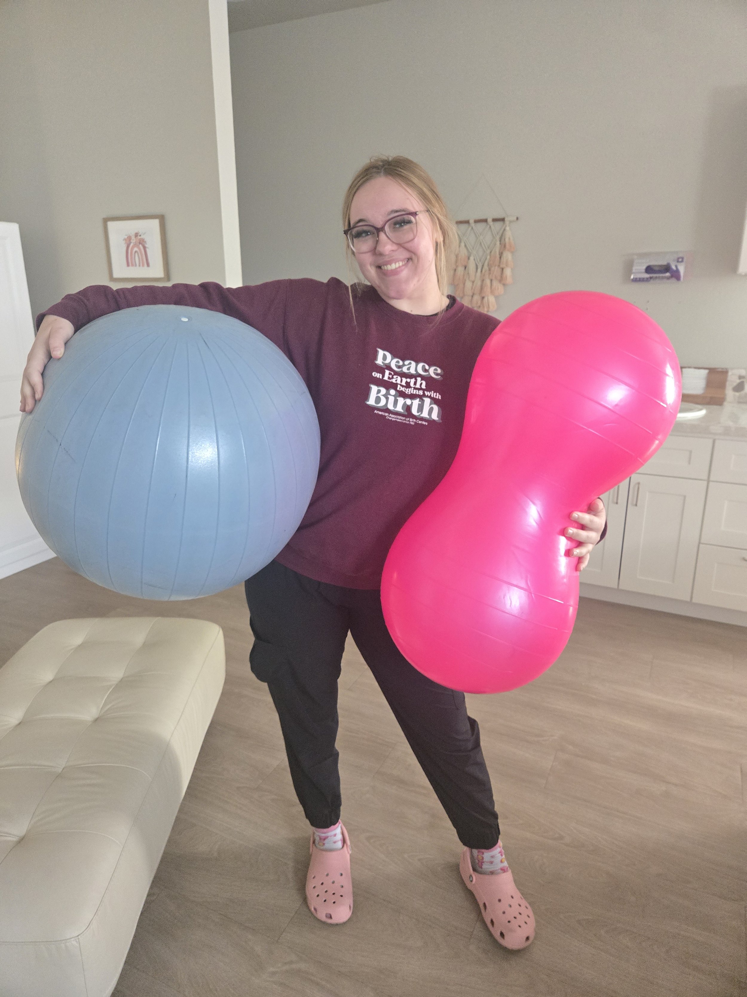 A woman with glasses smiling, wearing a maroon shirt with white text, holding a large blue exercise ball and a large pink heart-shaped balloon, standing in a room with a cream-colored couch, wooden floor, and wall decorations.
