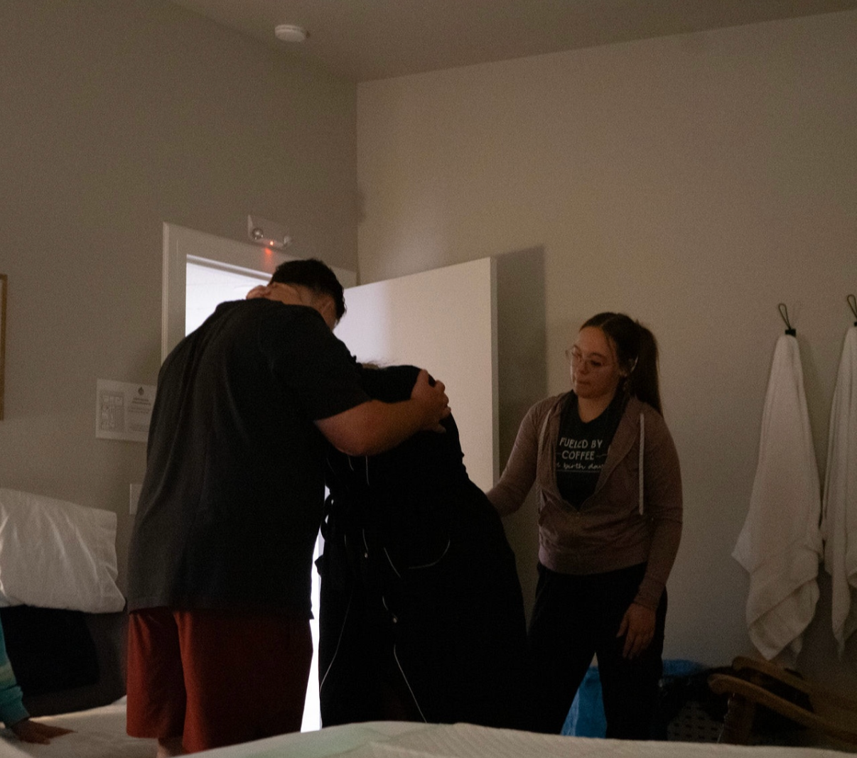 Three people gathered in a dimly lit hotel room, engaged in a comforting hug.