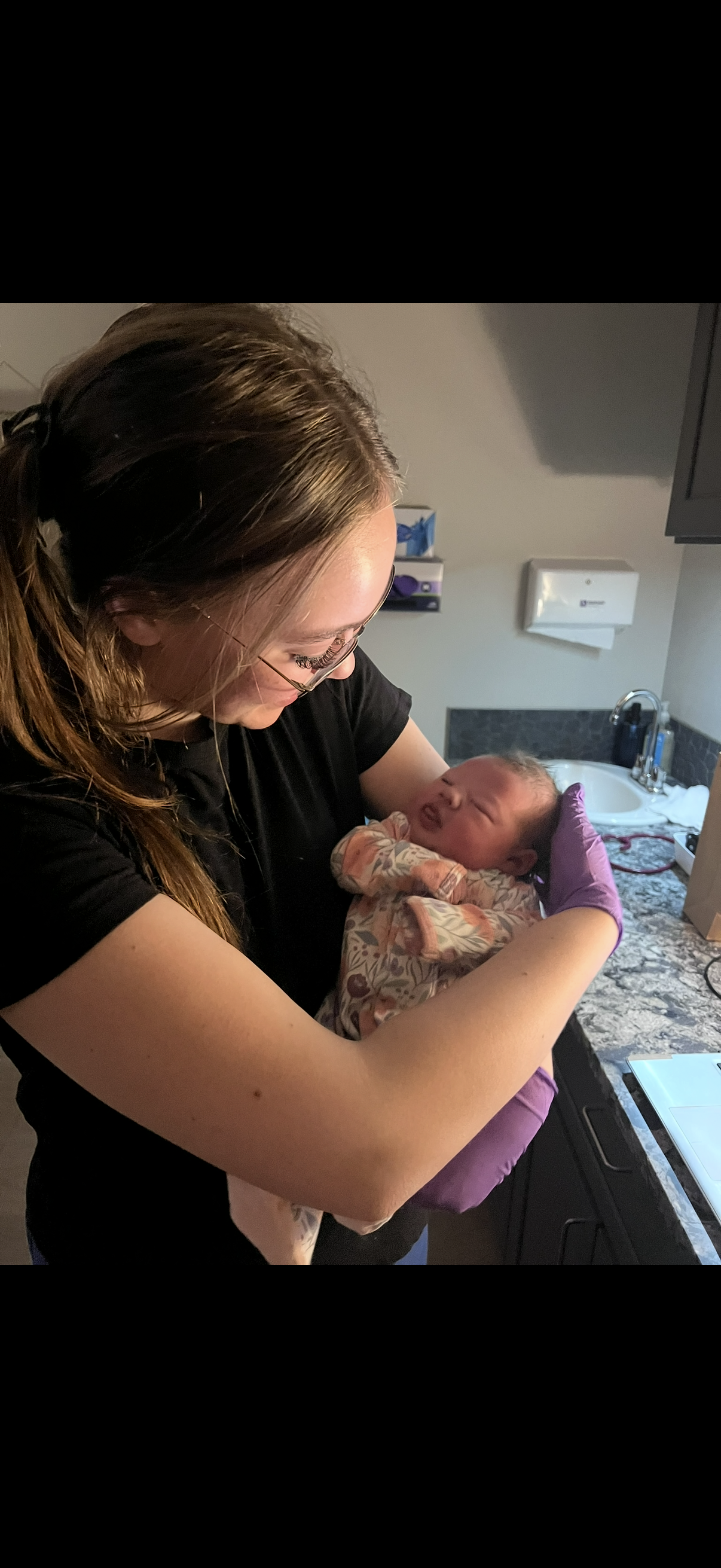 A woman holding a newborn baby in a hospital bathroom, smiling and looking down at the baby wrapped in a blanket.