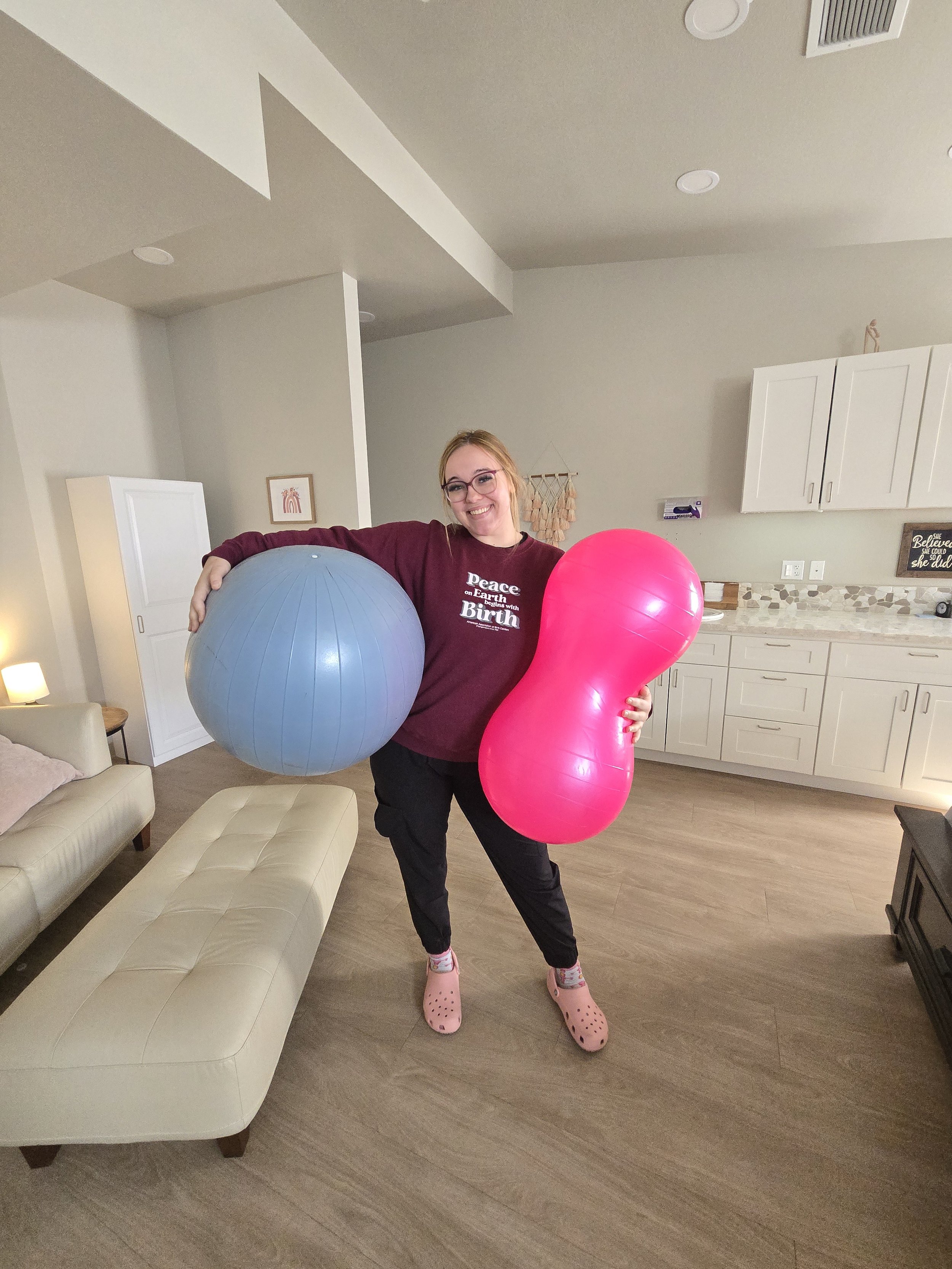 A woman with glasses and a maroon shirt smiling and holding two large balloons, one blue and one pink, in a modern kitchen and living room area.