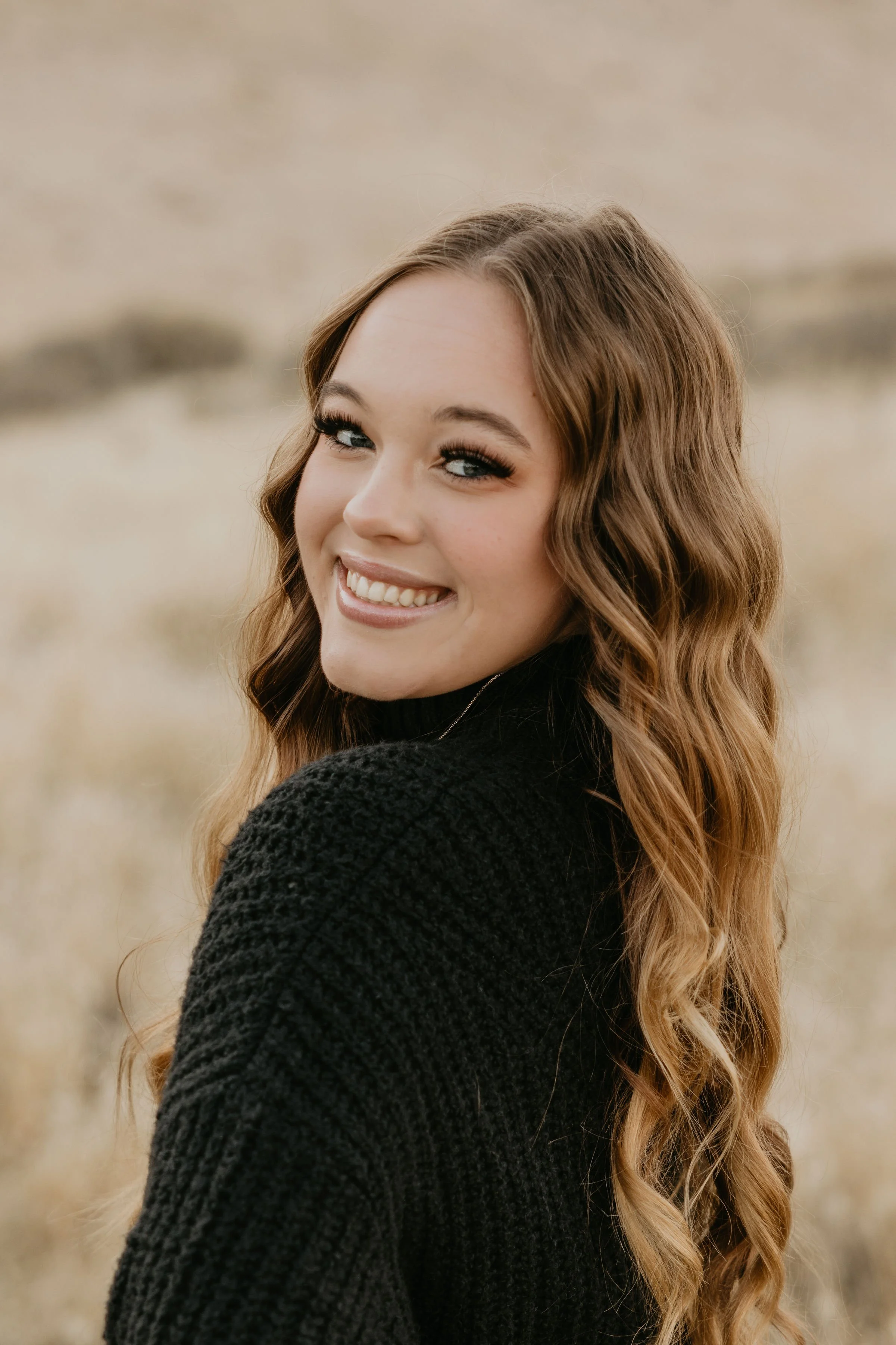 A young woman with long, wavy, light brown hair, smiling and looking at the camera. She is wearing a black sweater, with a blurred natural landscape background.