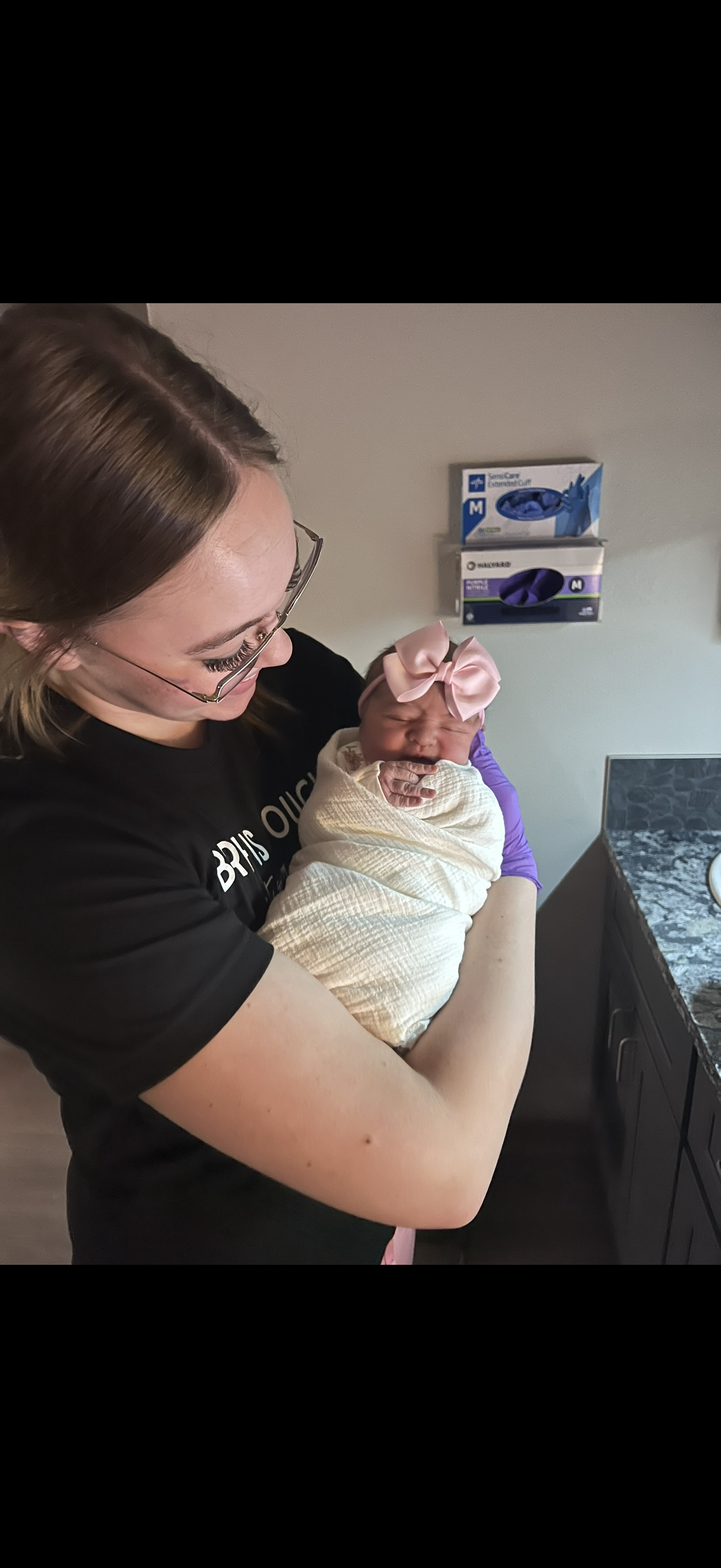 A woman holding a newborn baby wrapped in a blanket, with a pink bow headband, inside a room with medical supplies on the wall and a granite countertop.