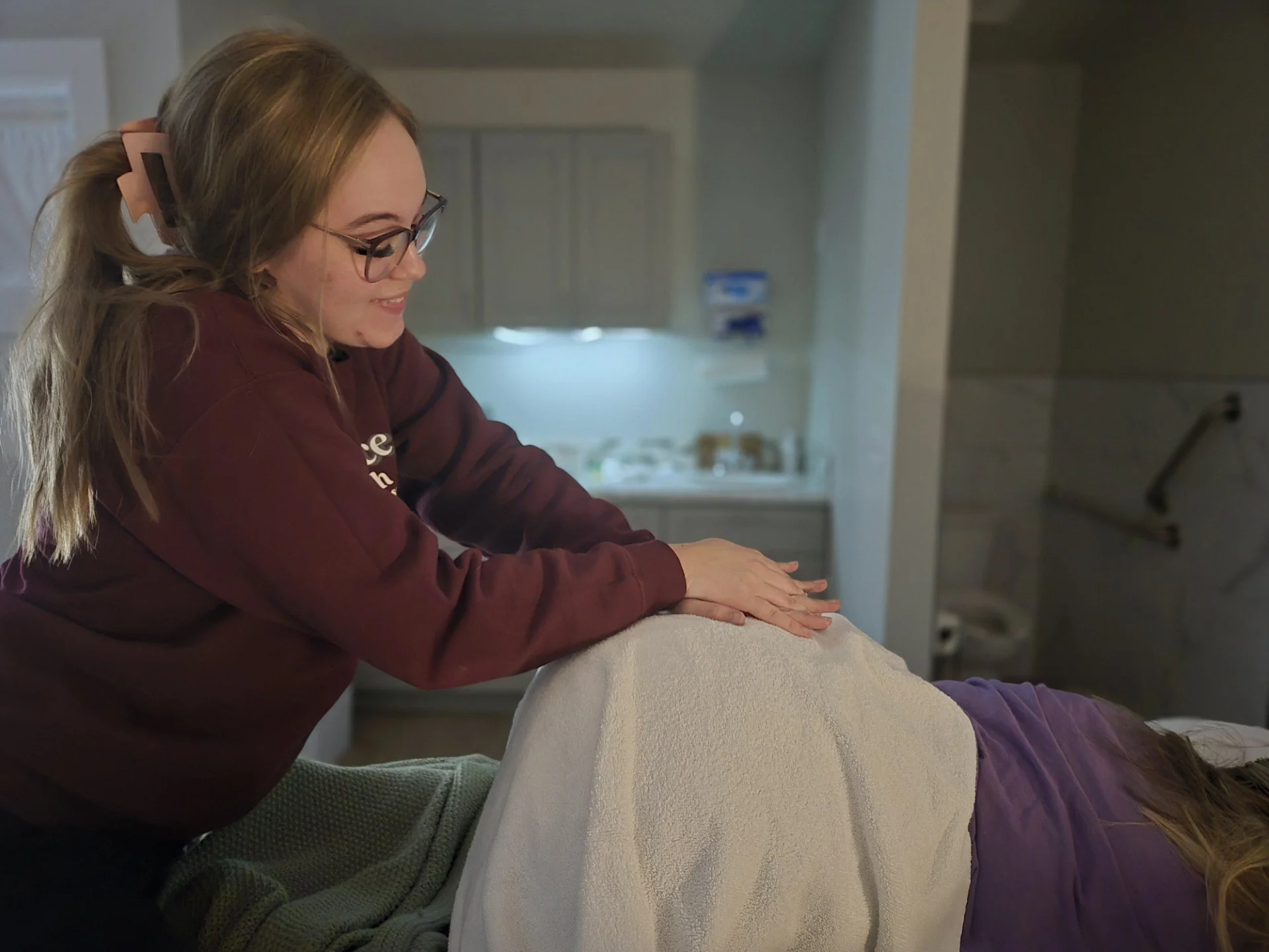 A woman with glasses and a hair clip is performing a massage or therapy on another person lying face down on a massage table.