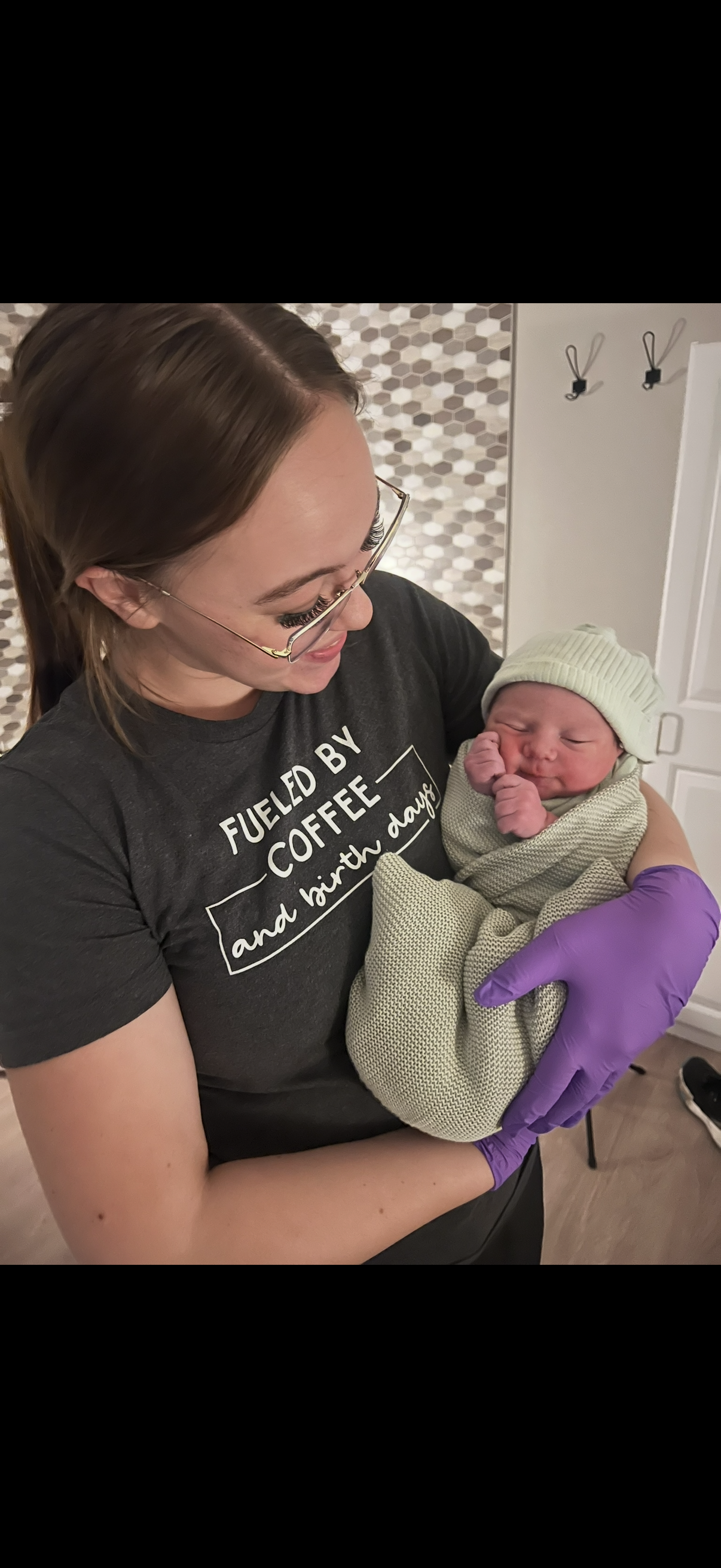 A woman holding a newborn baby, both smiling. The woman is wearing glasses, a dark T-shirt with the text 'Fueled by Coffee and Birth Days,' and purple gloves. The baby is wrapped in a green blanket and wearing a matching green hat, snuggling comfortably.