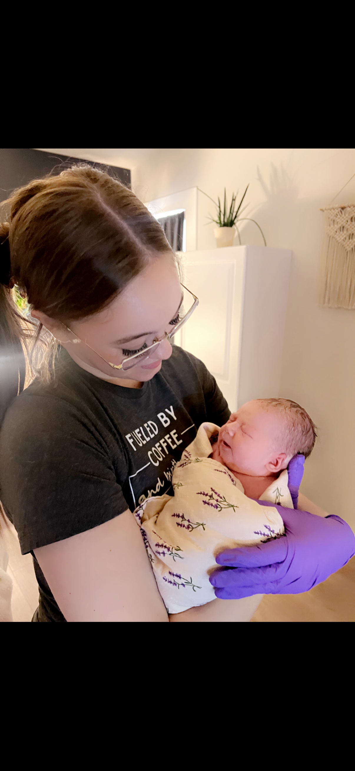 A woman with glasses and purple gloves holding a newborn baby wrapped in a floral blanket, smiling and looking at the baby in a cozy indoor setting.