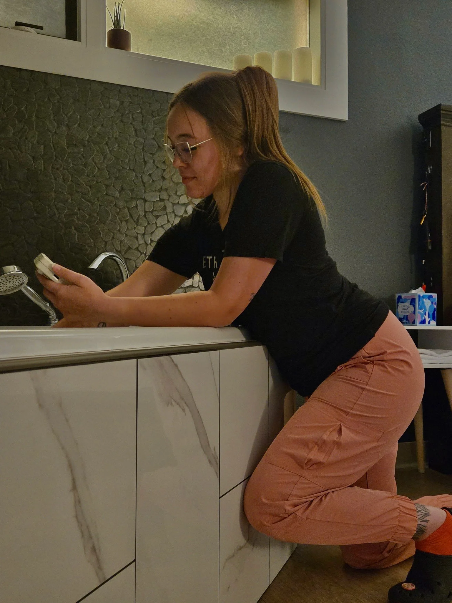 A woman with long hair in a ponytail, wearing glasses, a black t-shirt, and pink pants, is kneeling by a kitchen sink while holding a mobile phone.