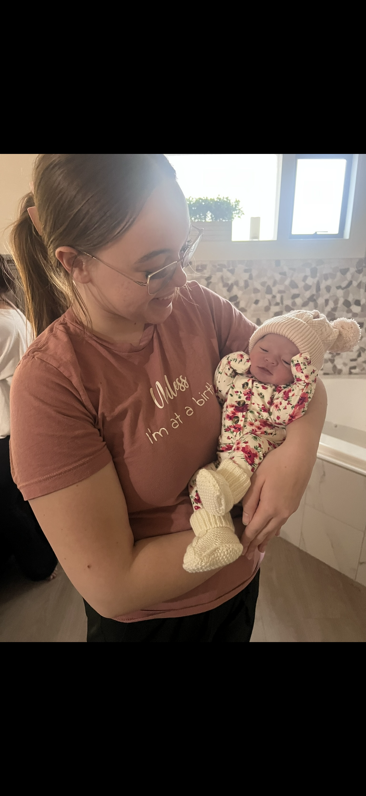A woman with glasses and a brown shirt holding a newborn baby dressed in a floral outfit with a knitted hat and booties. They are inside near a window.