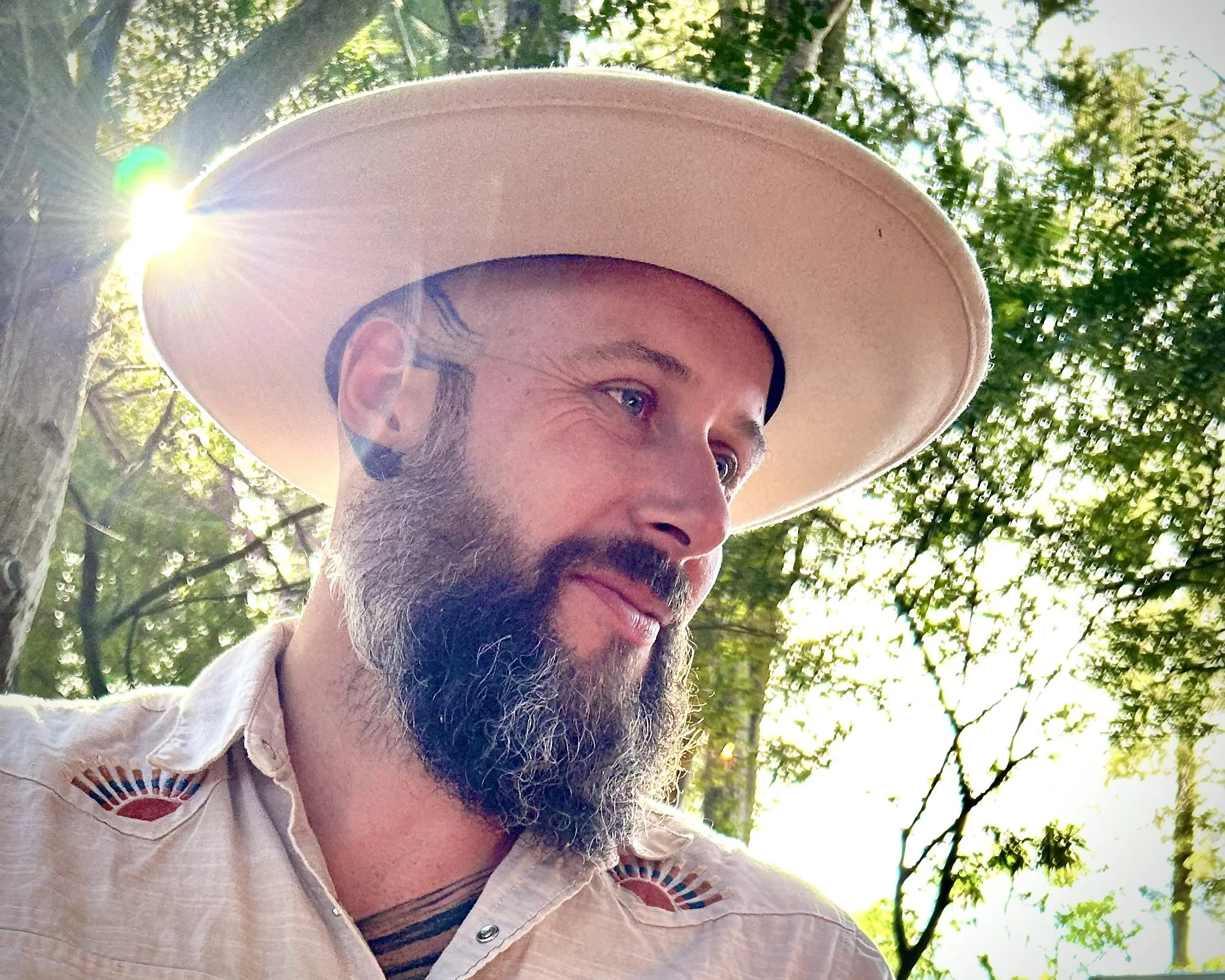A man with blue eyes and facial hair wearing a light-colored shirt with embroided suns on the shoulders, a beige cowboy hat, standing outdoors with sunlight filtering through green trees in the background.