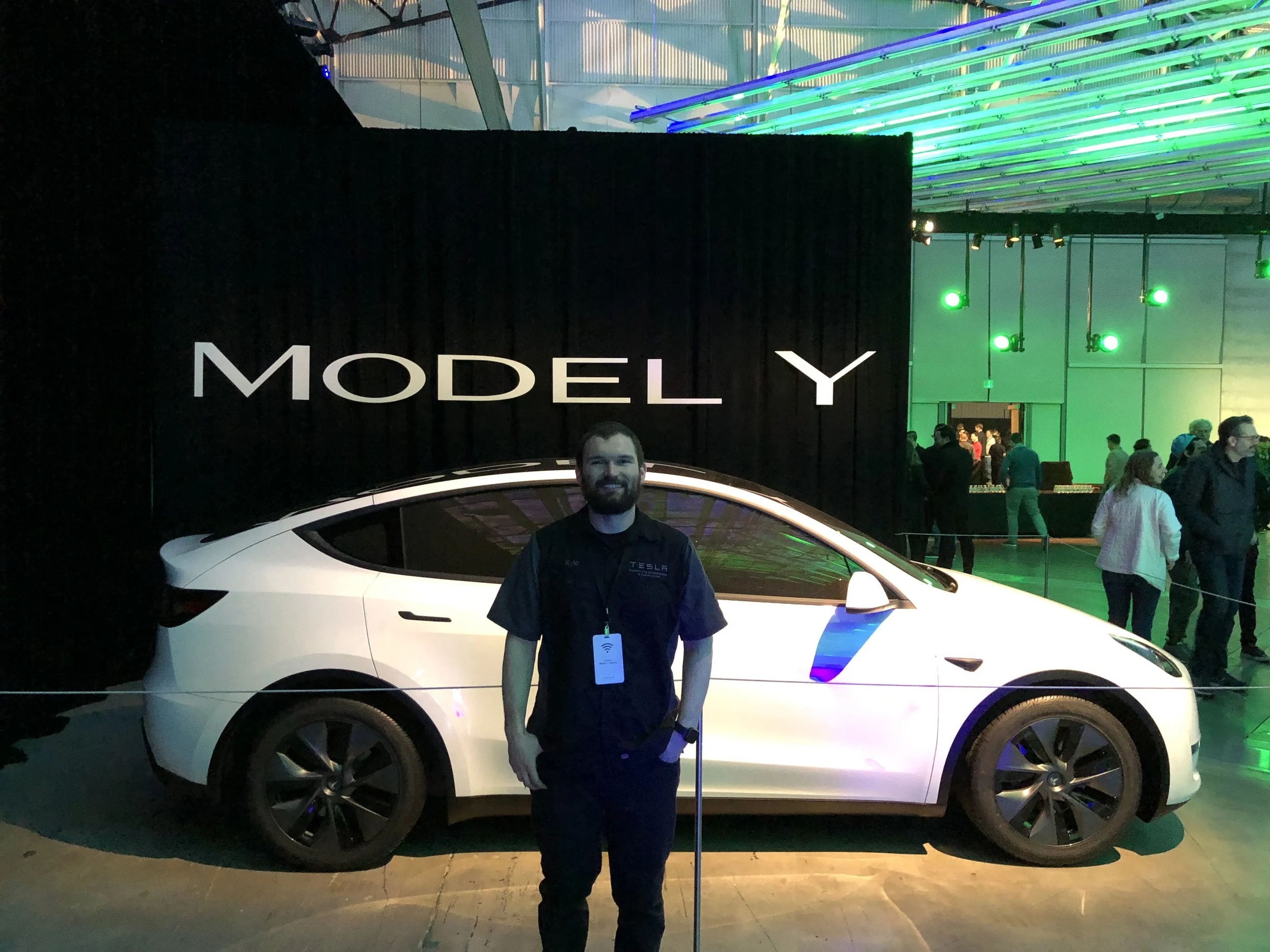 A man wearing a Tesla shirt standing in front of a white Tesla Model Y. The background shows an indoor event with bright green and blue lighting, a black curtain, and colorful neon lights on the ceiling. The sign in the background reads 'MODEL Y'.