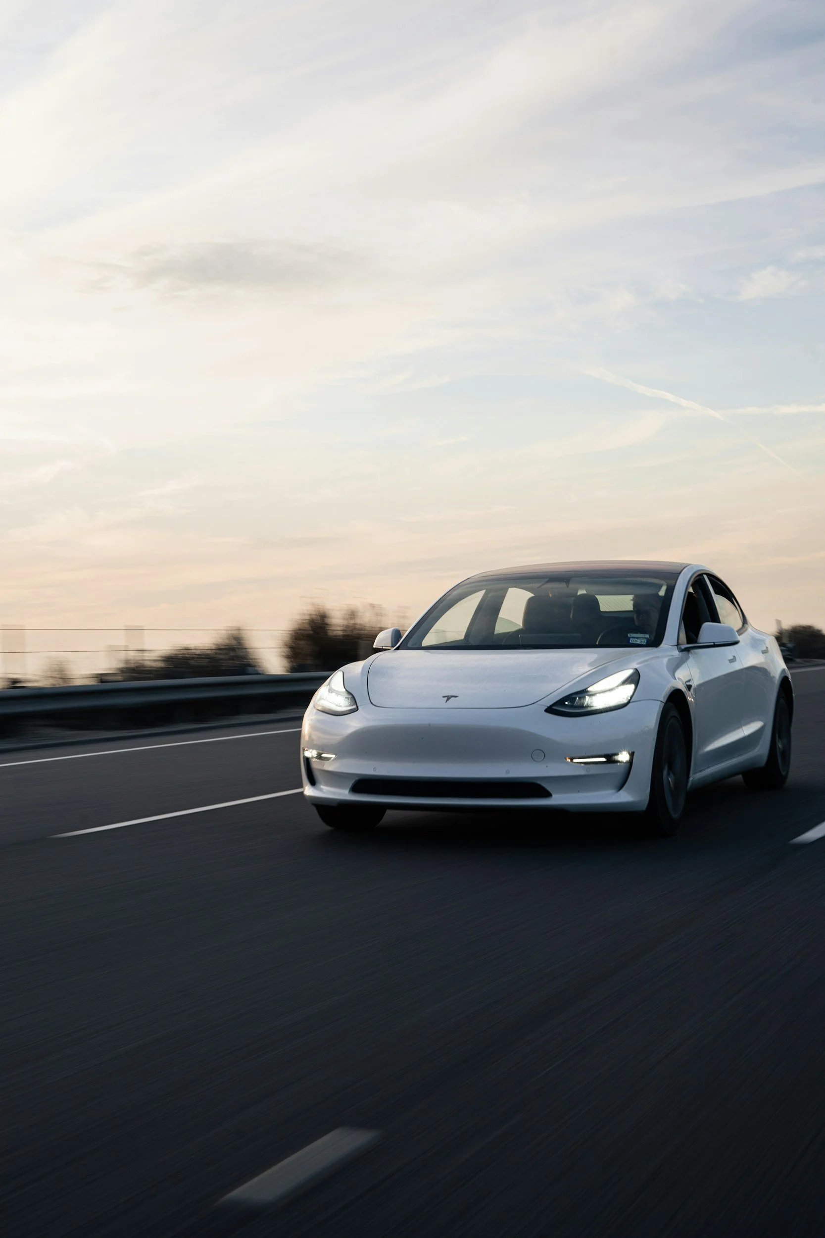 A white Tesla Model 3 driving on a highway during sunset or sunrise with a mostly cloudy sky.