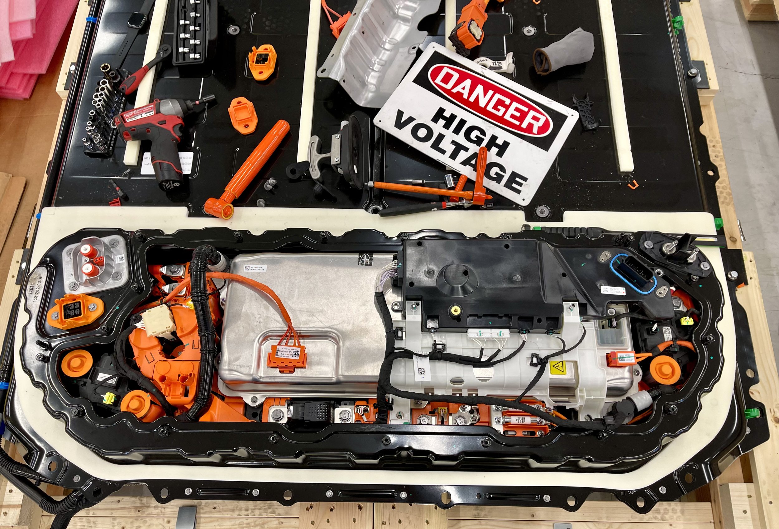 Interior of an electric vehicle's battery pack with various components, wiring, and electronic parts, alongside a warning sign that reads 'Danger High Voltage'.