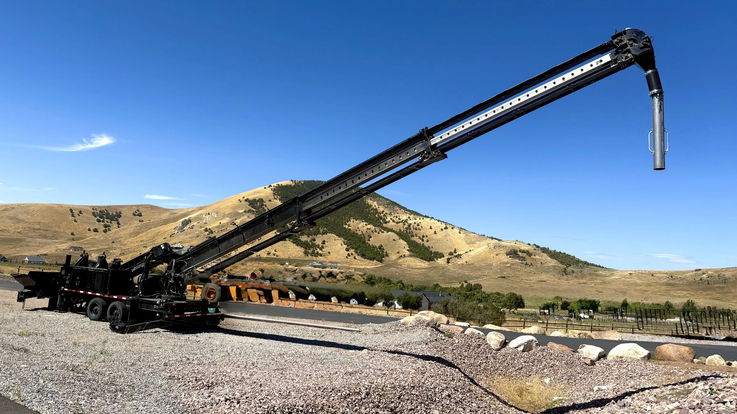 A large black crane truck working outdoors in a rural area with hills and houses in the background under a clear blue sky.