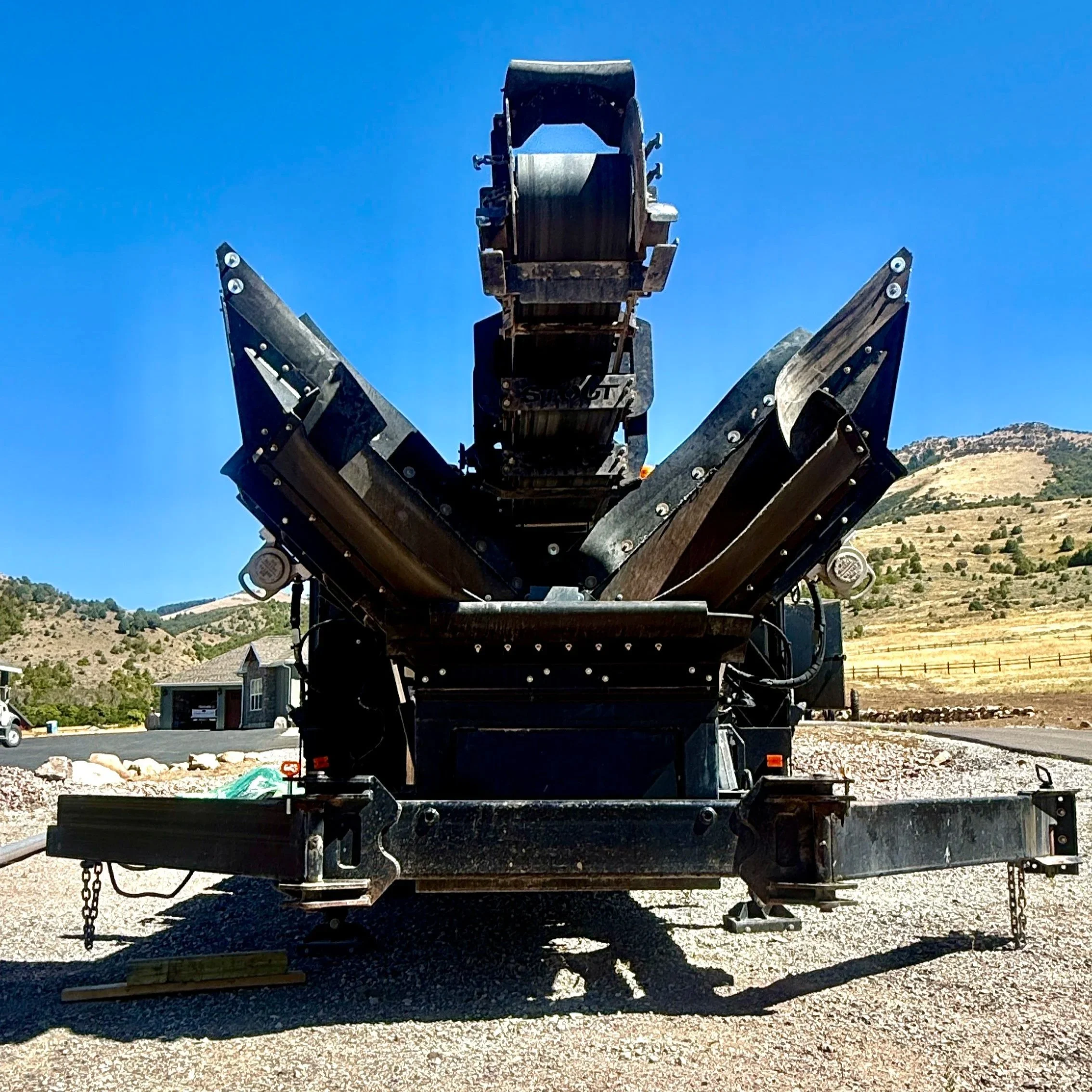 A large construction machine with a long, extendable arm and a bucket attachment, parked outdoors on gravel near a small building, with a mountainous landscape in the background.