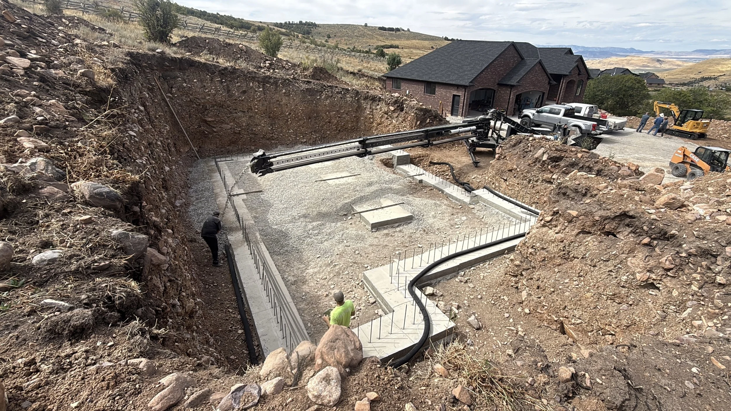 Construction workers work on a house foundation with excavation, gravel, and concrete. Heavy machinery and trucks are on site in a suburban area with hills in the background.