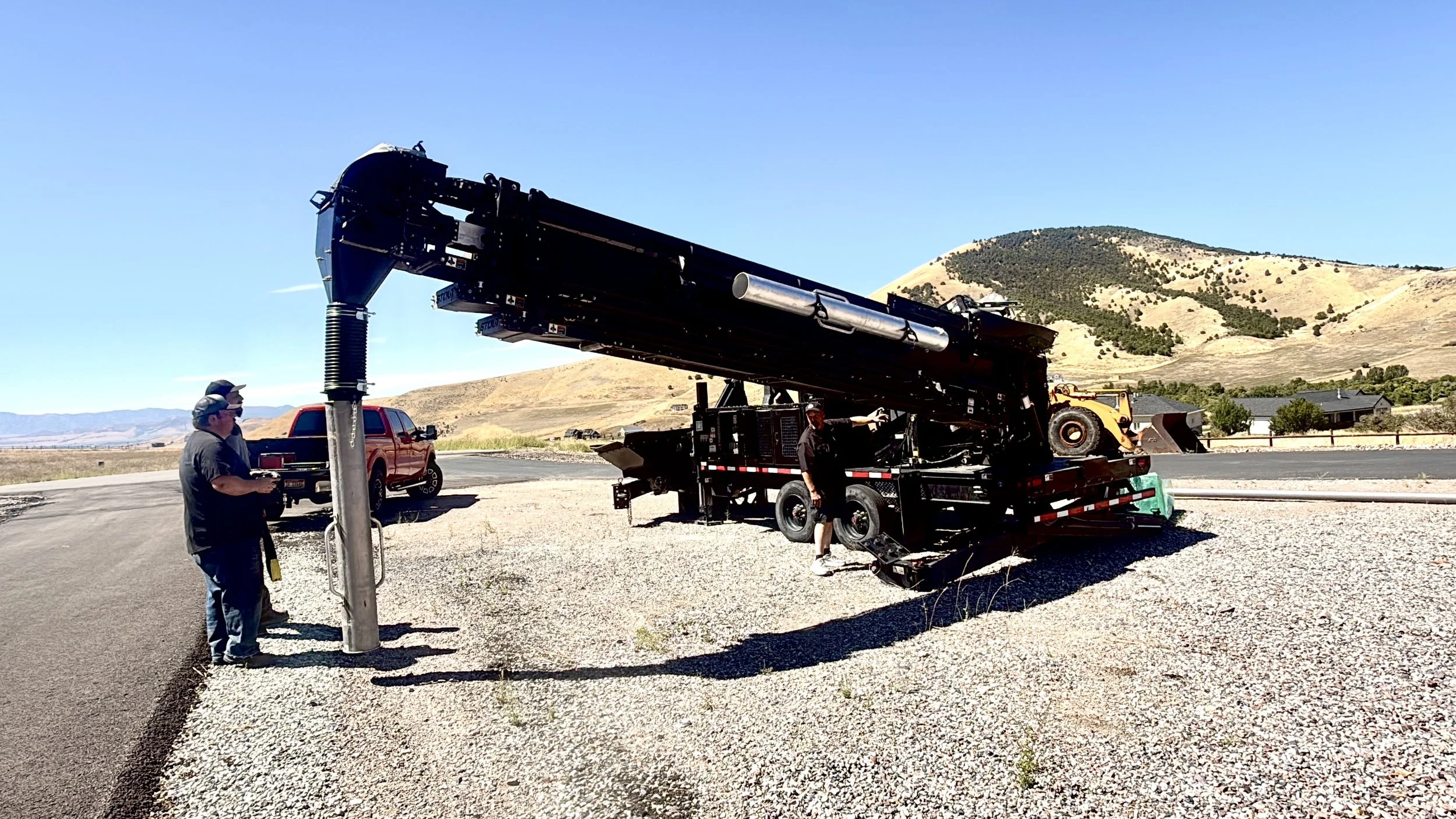 People working with a large piece of machinery on a trailer in a rural area with mountains in the background.