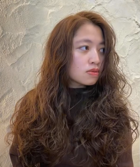 A woman with long, curly brown hair looking to her right, wearing a dark top, standing against a textured beige wall.