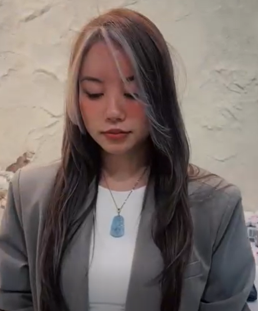 Young woman with long, dark hair and blonde highlights wearing a gray blazer and white shirt, looking down with a neutral expression, standing against a textured light-colored wall.