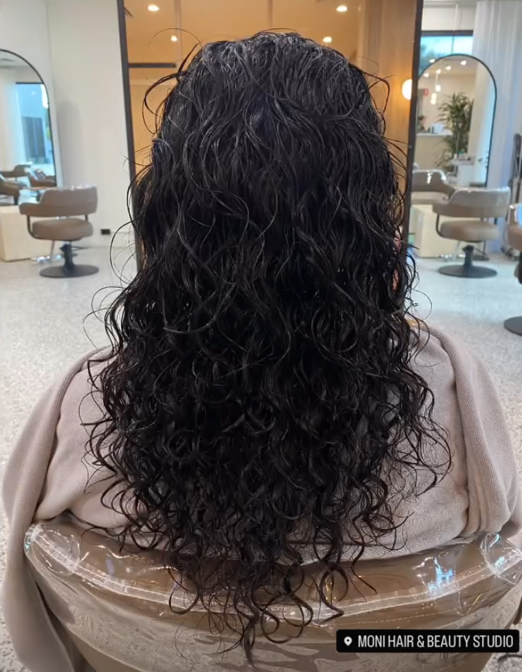 Back view of a person with long, curly, dark hair sitting in a salon chair. The salon interior in the background features beige salon chairs, mirrors, and bright lighting.