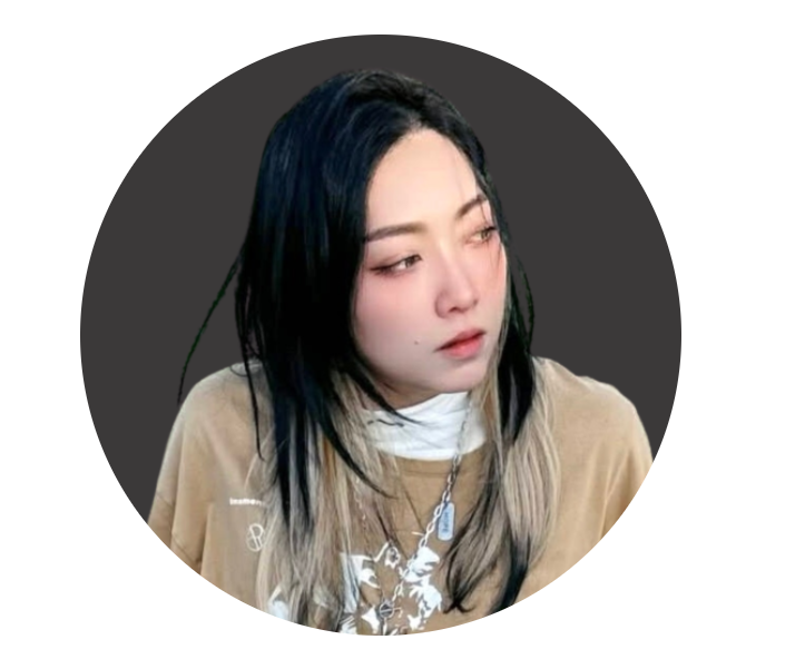 A young woman with black and blonde hair looking to the side with a neutral expression, wearing a beige shirt and layered necklaces, against a dark round background.