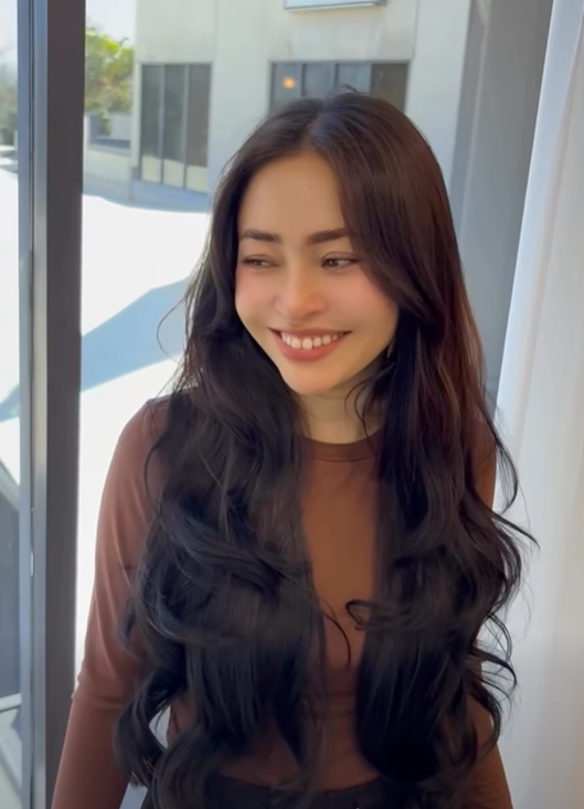 Young woman with long wavy dark hair smiling, standing in front of a window.